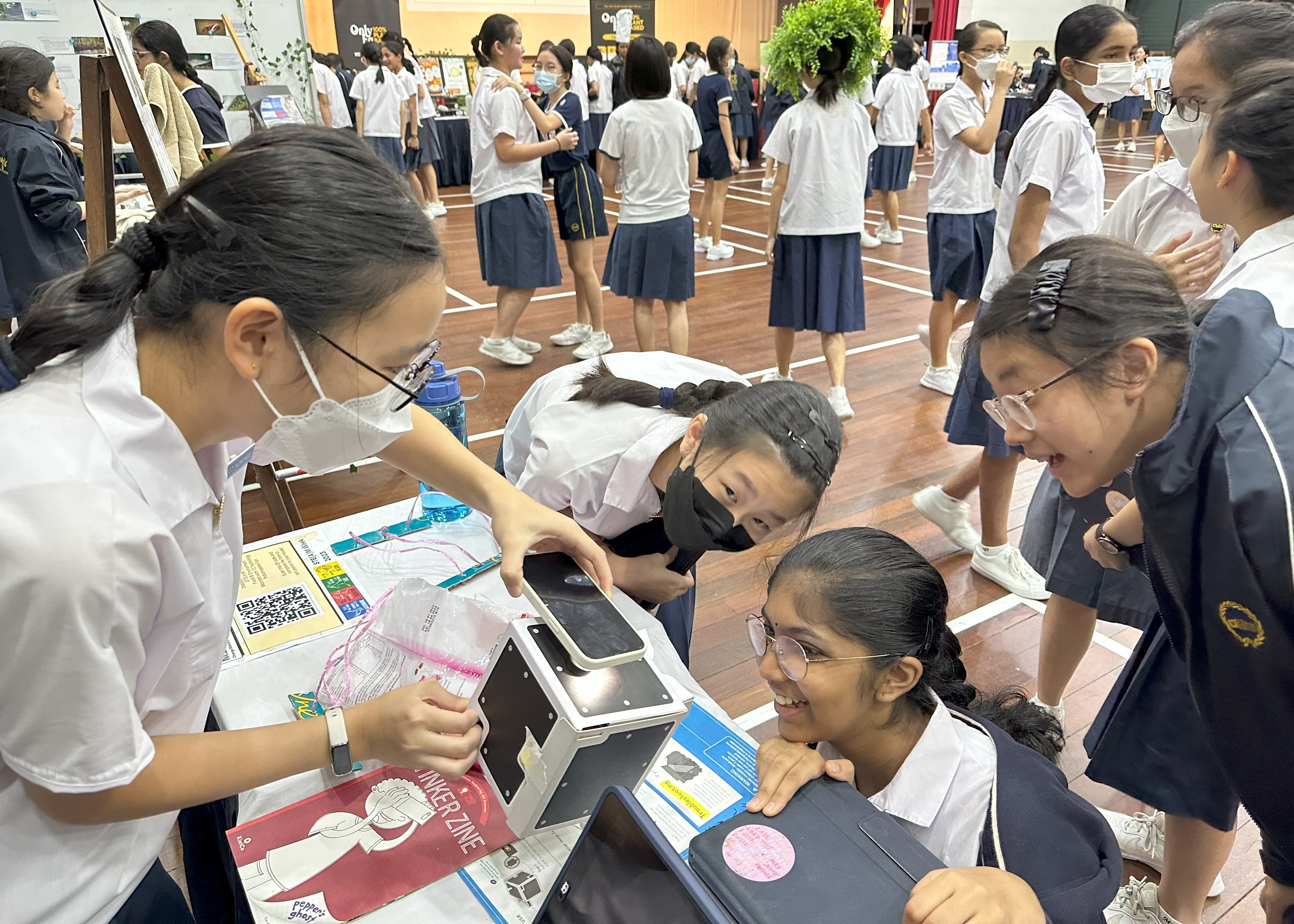 School girls in uniforms gather around a table with a device during what appears to be a science fair in a hall.