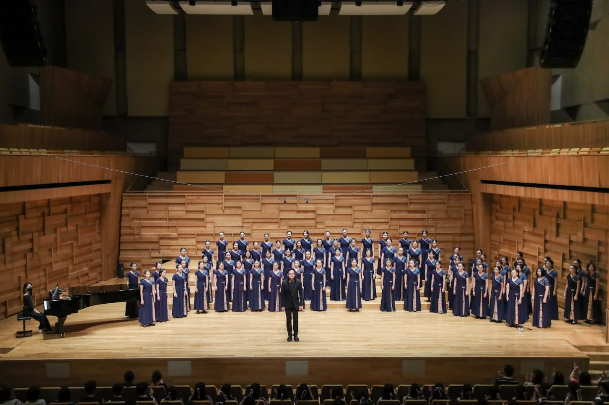 A large choir in blue gowns stands on a concert hall stage with a conductor in black and a pianist.