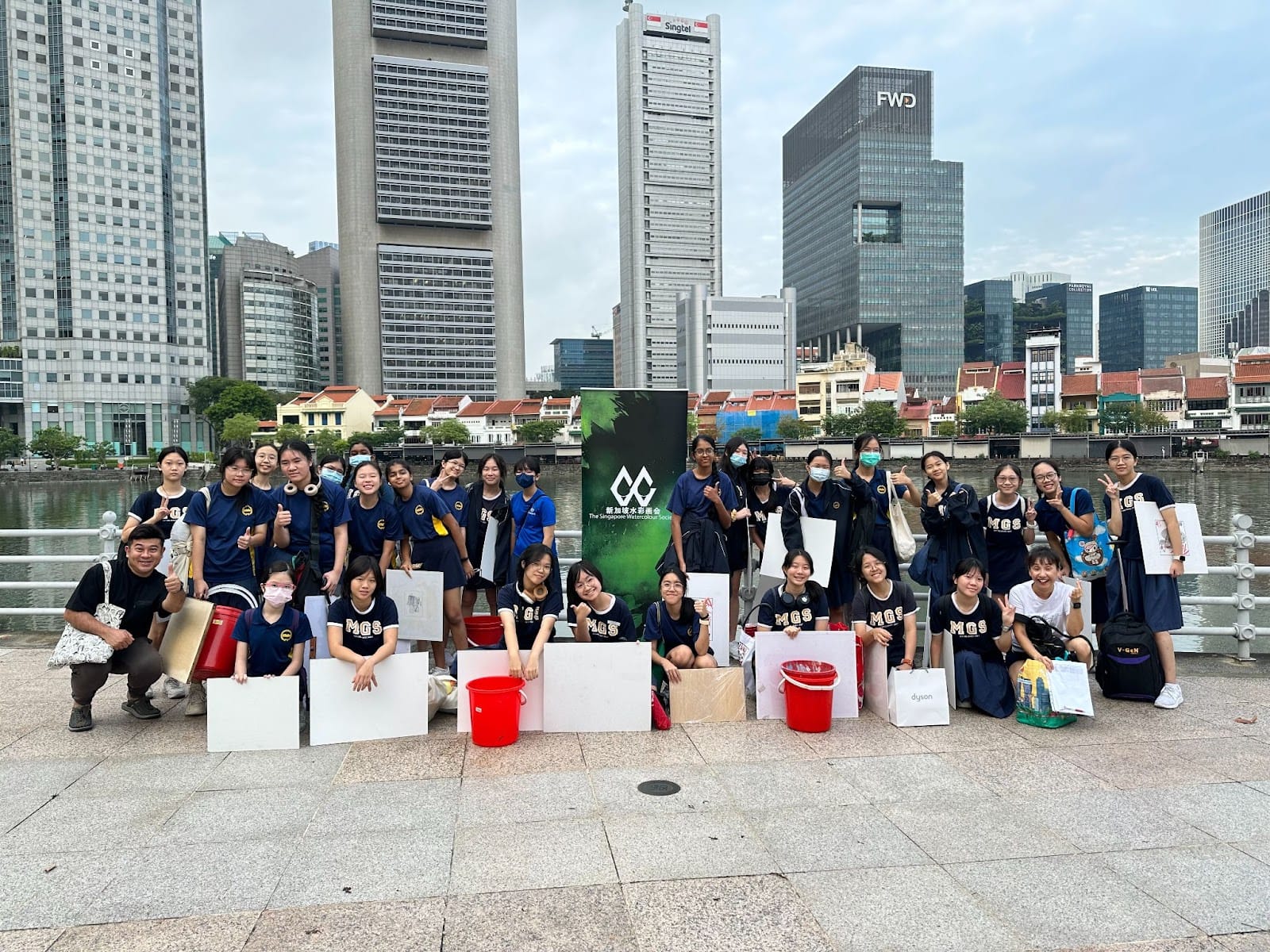 Group of students in navy MGS uniforms pose with art boards and buckets by a Singapore river with city skyscrapers behind them.