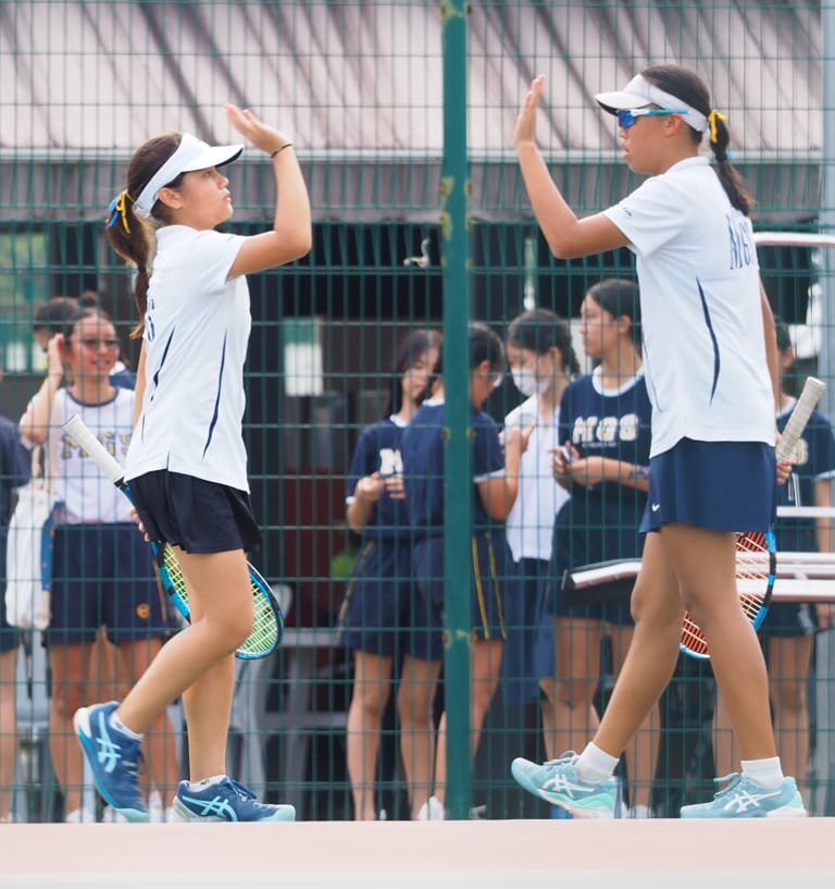 Two young female tennis players in white uniforms high-fiving on a court with teammates watching behind a fence.