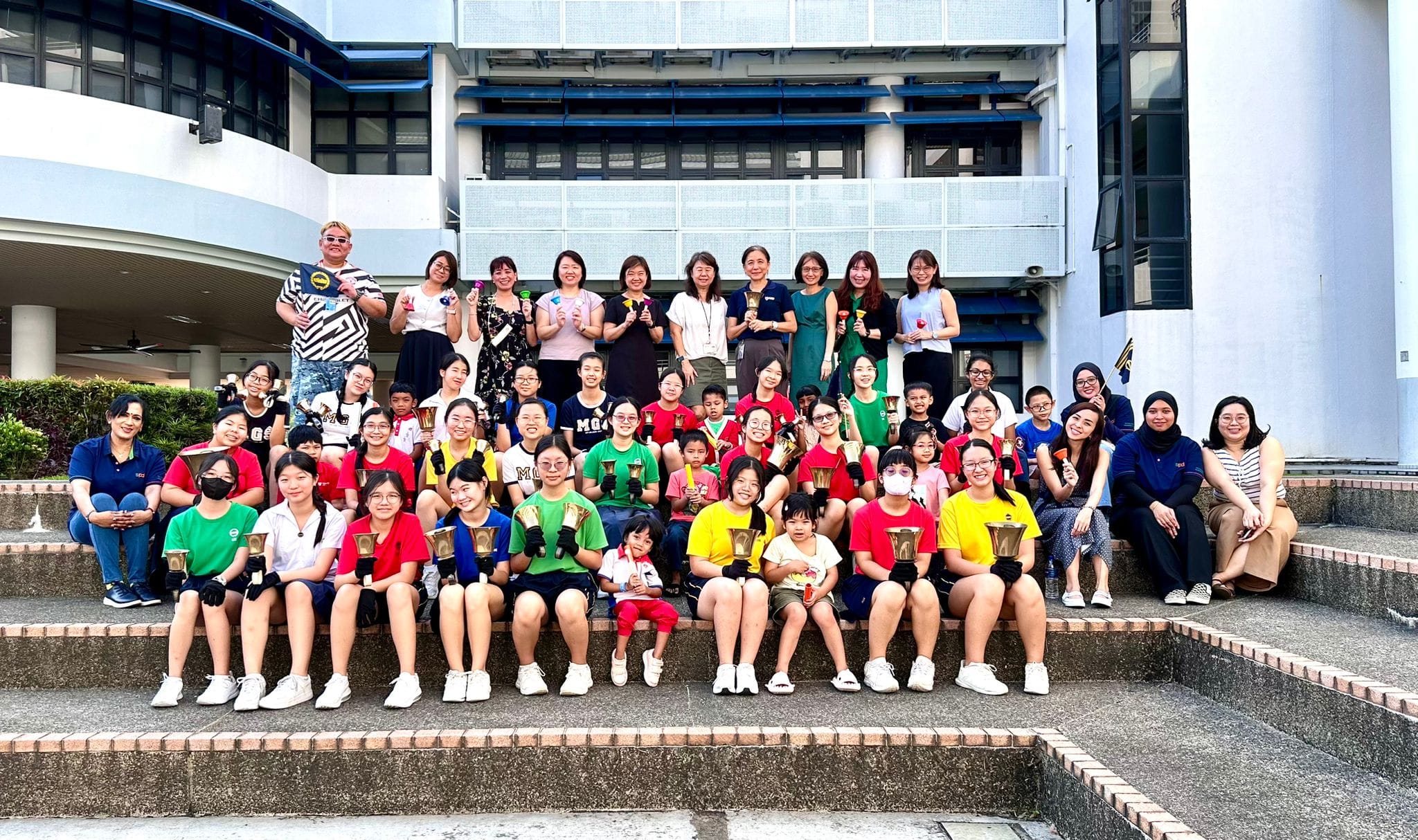 A large group of students and adults pose on outdoor steps, many holding handbells, in front of a school building.