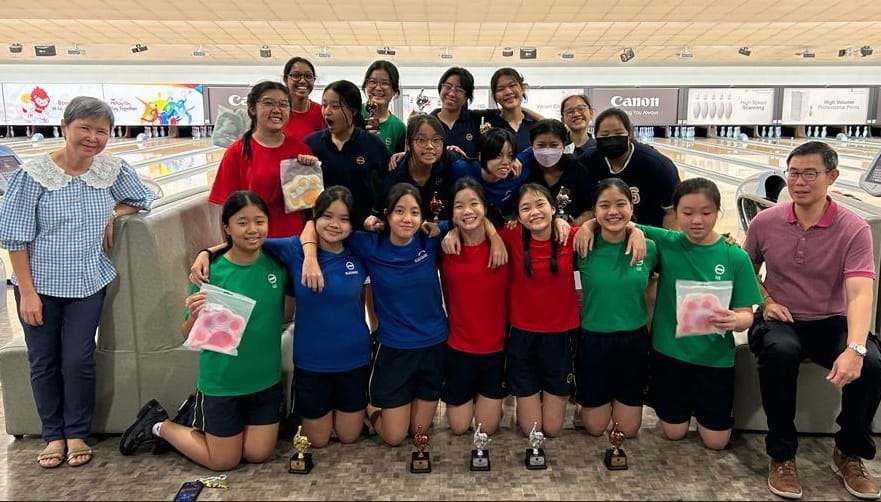 A group of young female bowlers pose with trophies at a bowling alley, joined by two adult coaches.