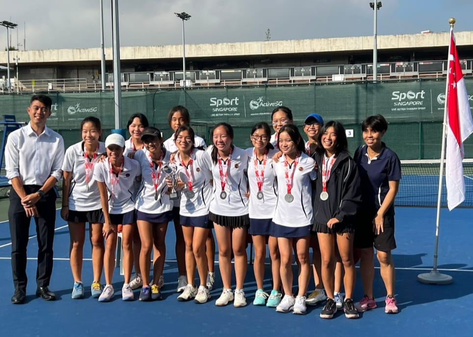 A group of young female tennis players wearing medals pose with a trophy on a Sport Singapore court.