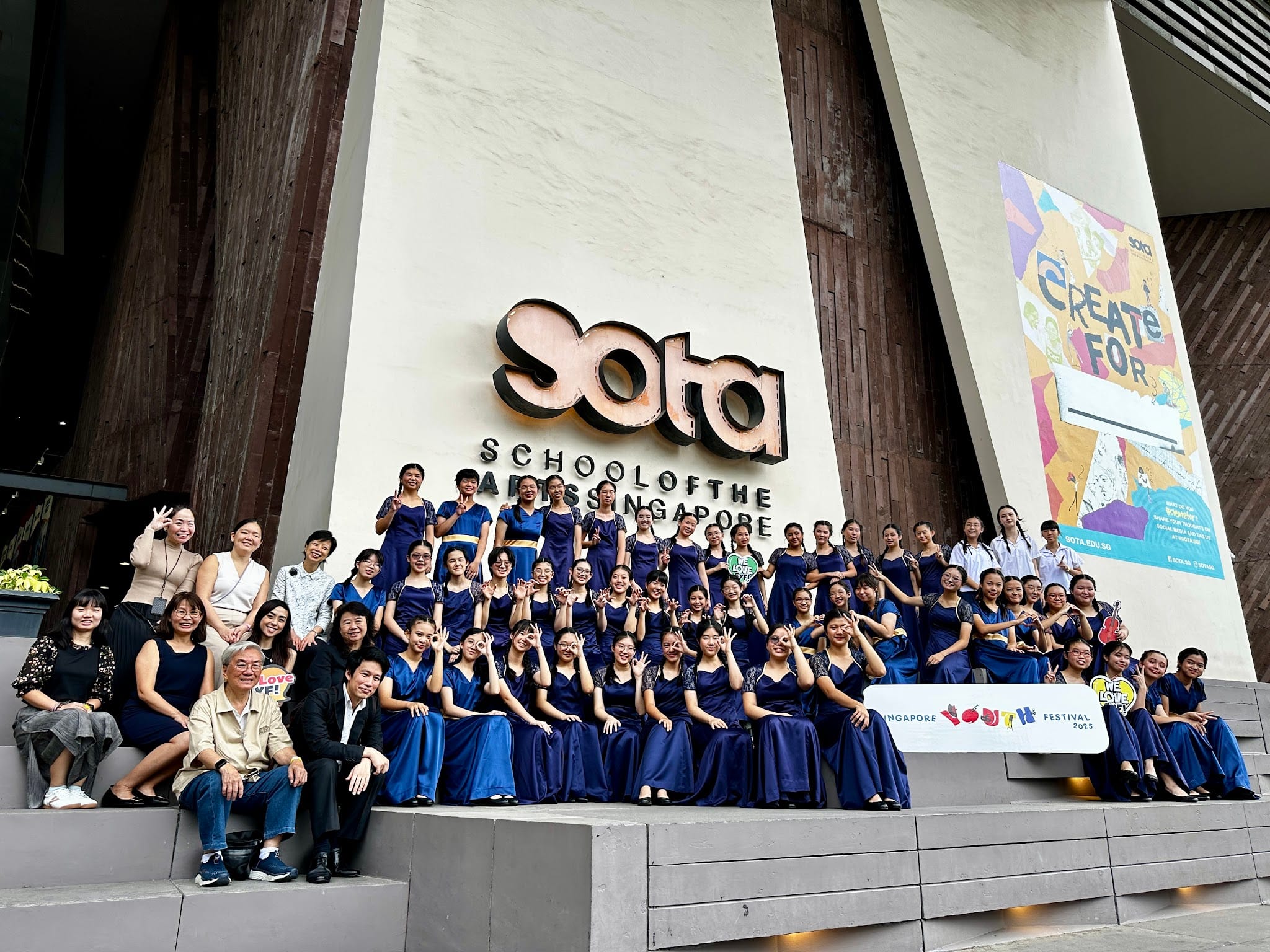 Large group in navy blue gowns posing on steps in front of the SOTA School of the Arts Singapore building.