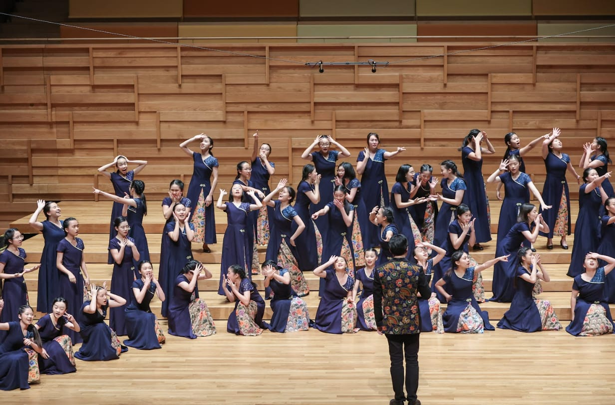 A large choir of girls in navy dresses performs on a wooden stage, led by a conductor facing them.