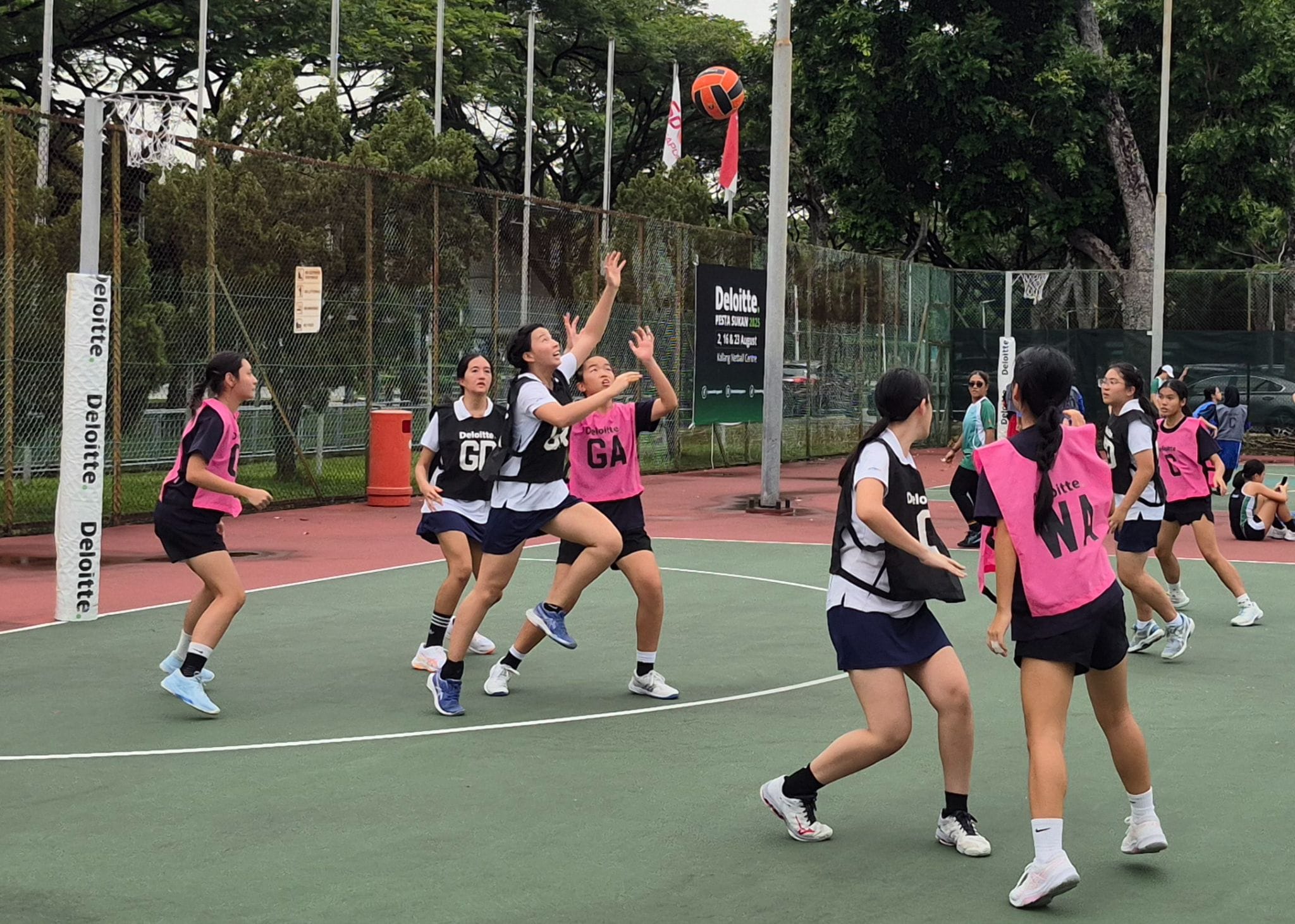 Girls in pink and dark bibs playing netball on an outdoor court, with a ball in the air near the goal post.