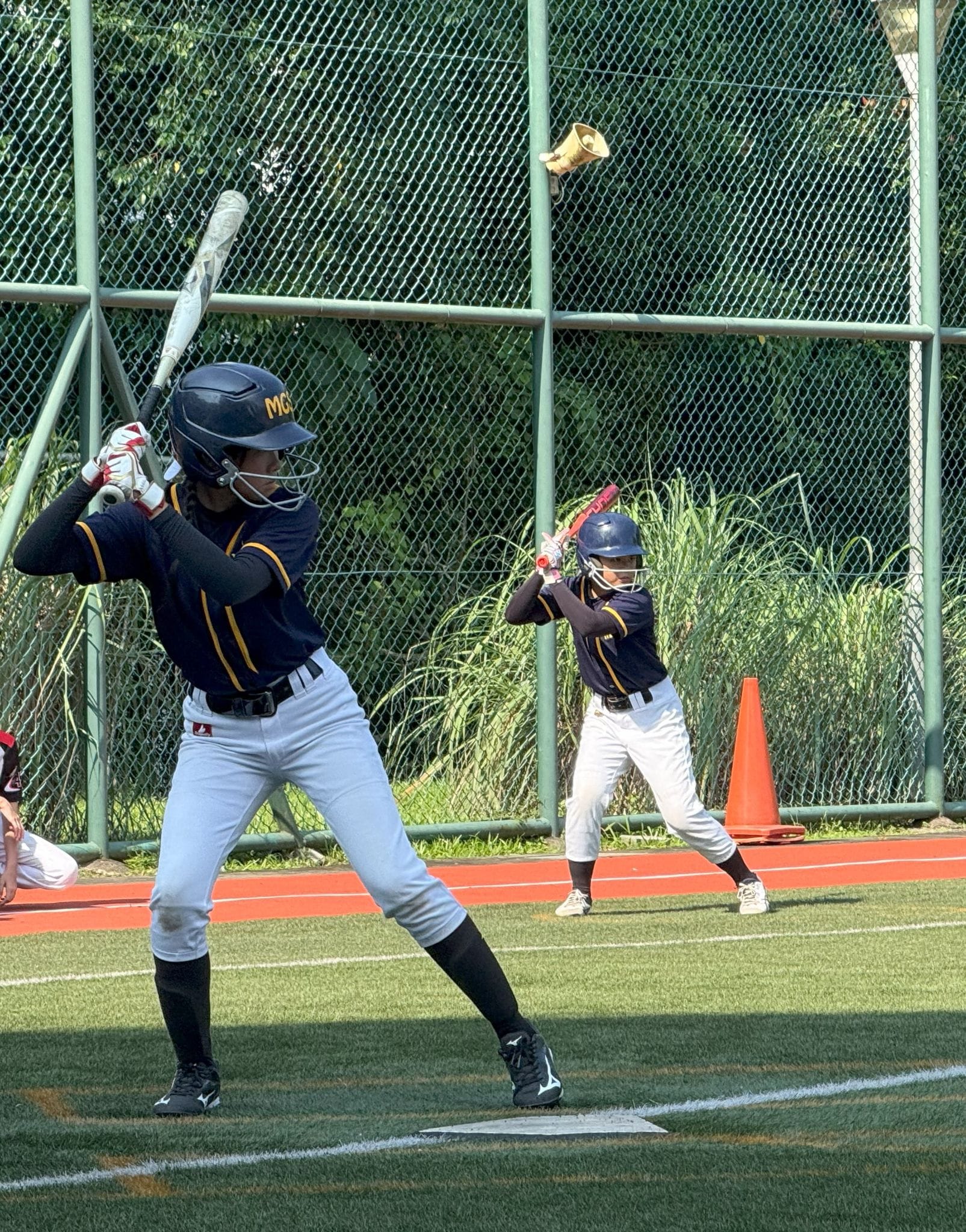 Two youth softball players in navy uniforms and helmets stand in batting stance on a green turf field.