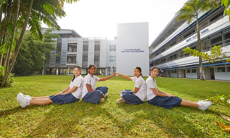 Four schoolgirls in white uniforms sit on grass in front of the Puan Sri Helene Tan Chin Tuan Building.