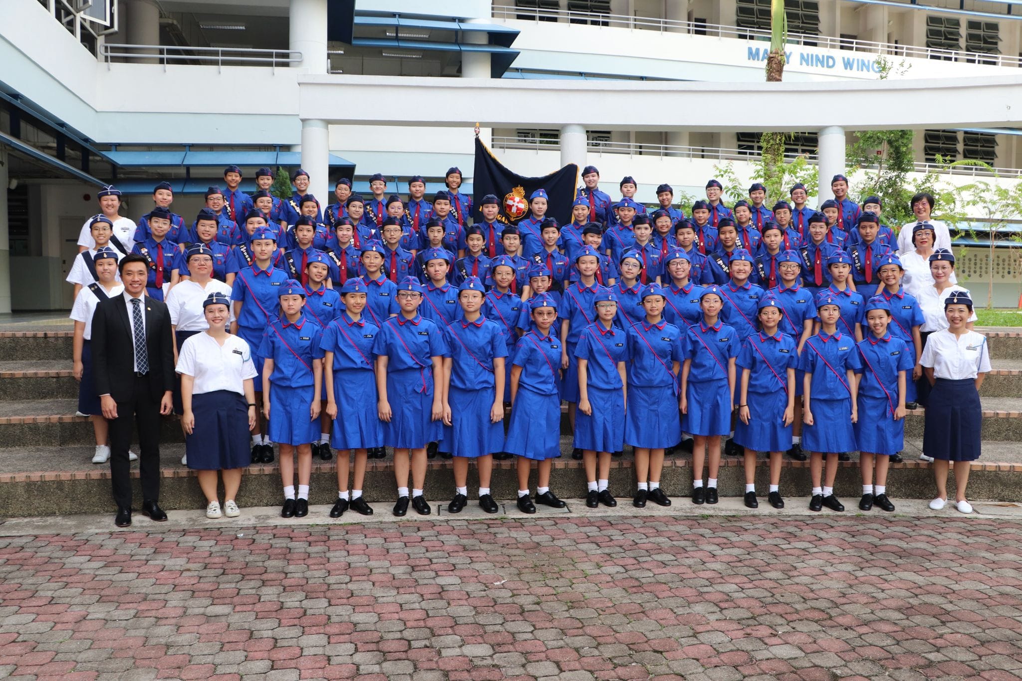 Large group of students in blue uniforms and caps posing for a formal photo outside a school building.