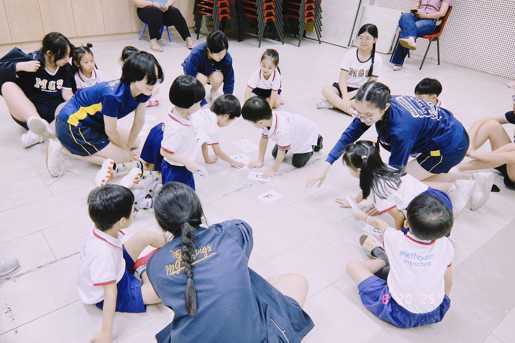 Young children in preschool uniforms sit on a floor in a circle with older students arranging letter cards.