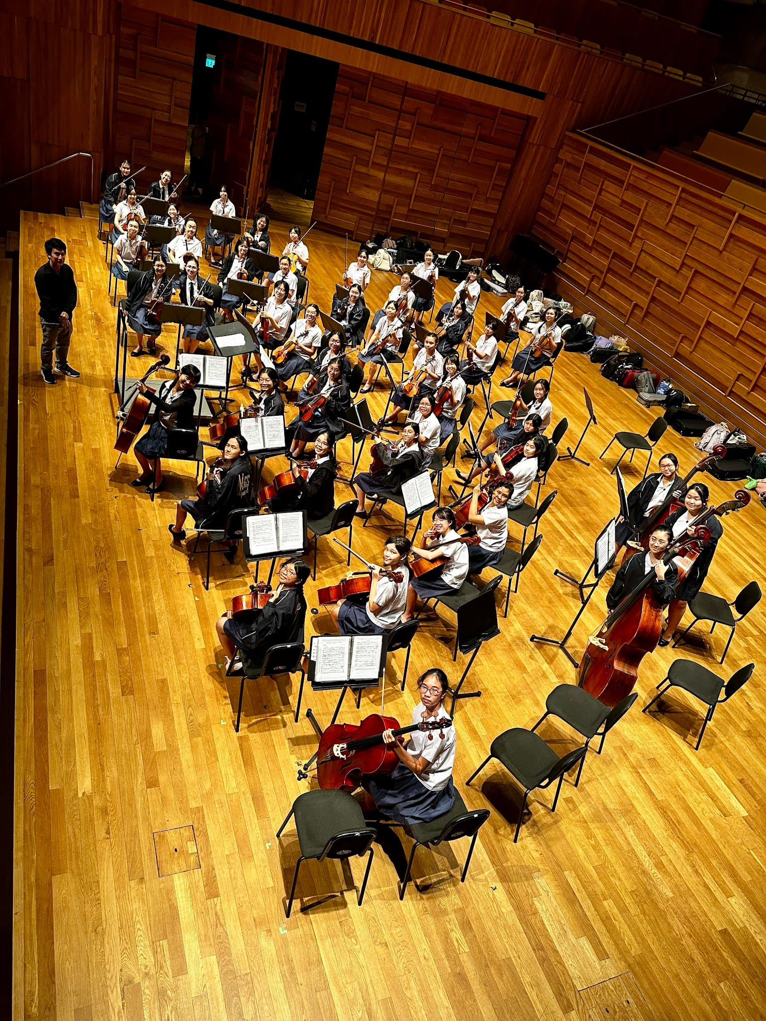 A large youth string orchestra seated on a wooden concert hall stage with music stands, holding violins and cellos.