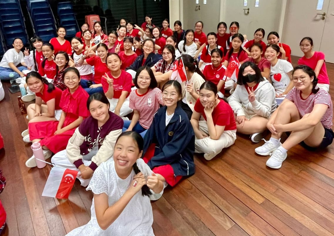 Large group of smiling girls in red and white outfits holding Singapore flags, seated on a wooden floor.