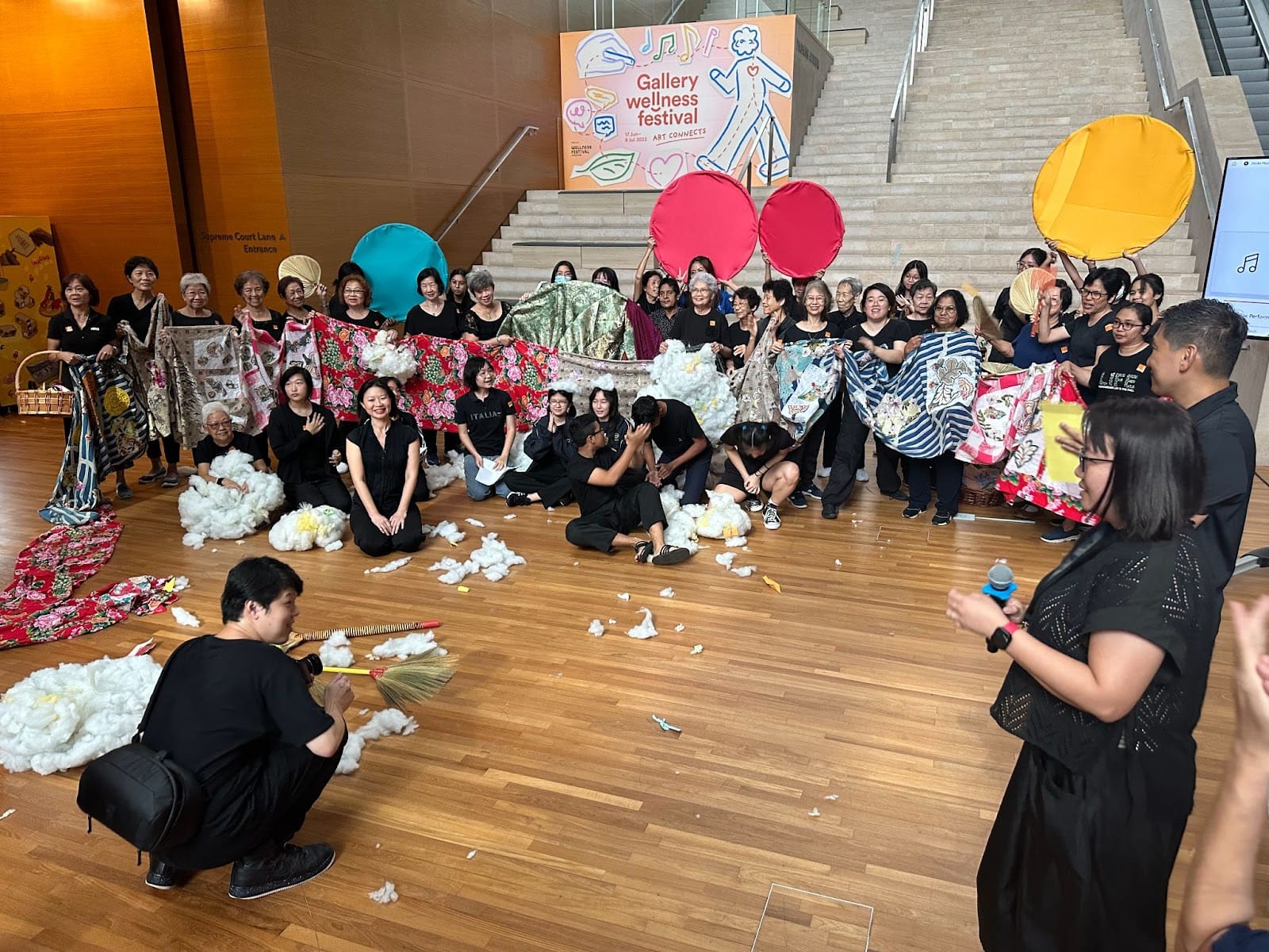 A large group in black clothing holds colorful fabrics and round props at a Gallery Wellness Festival event indoors.