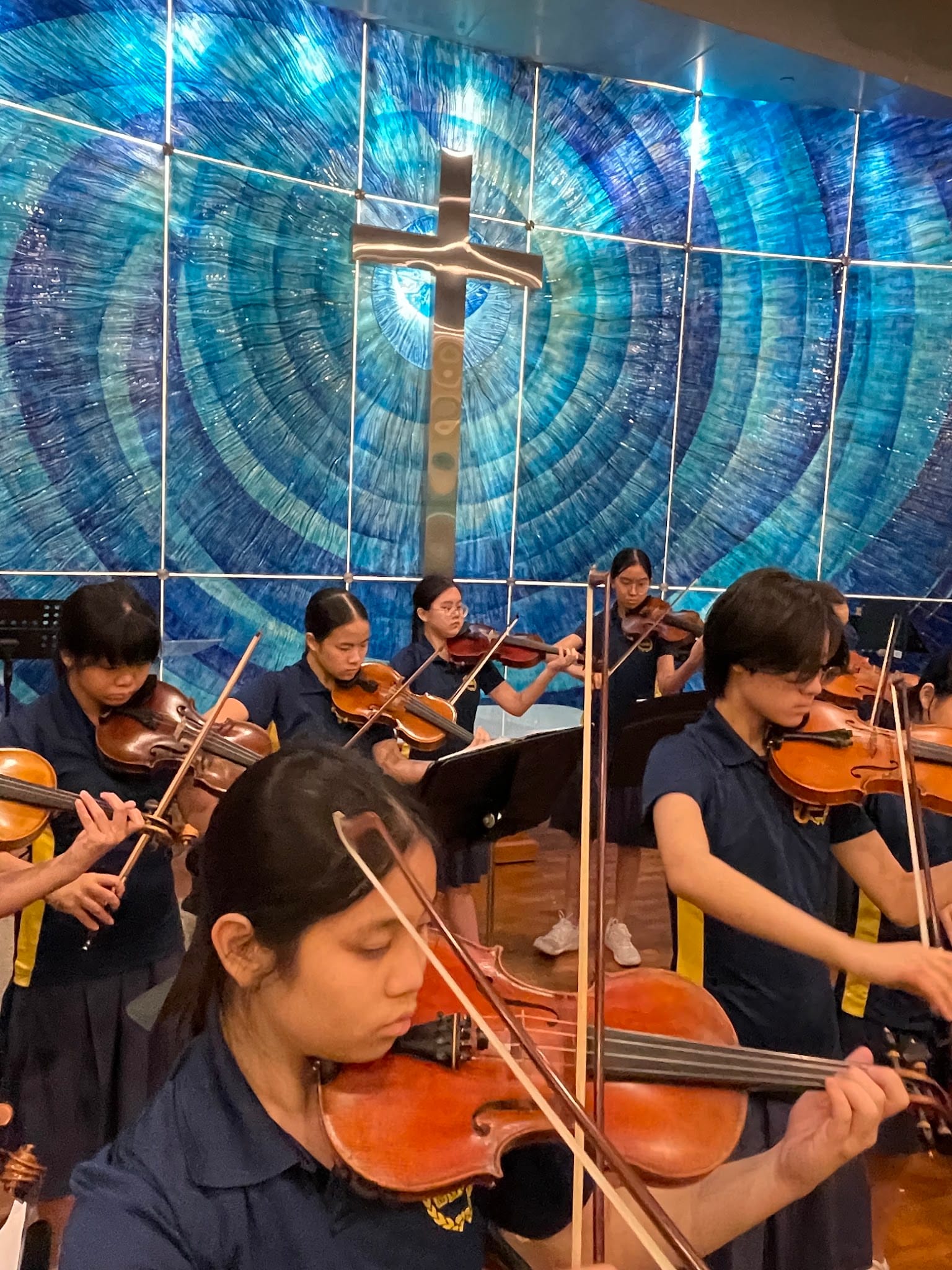 Young students in navy uniforms playing violins in front of a blue stained glass wall with a cross.