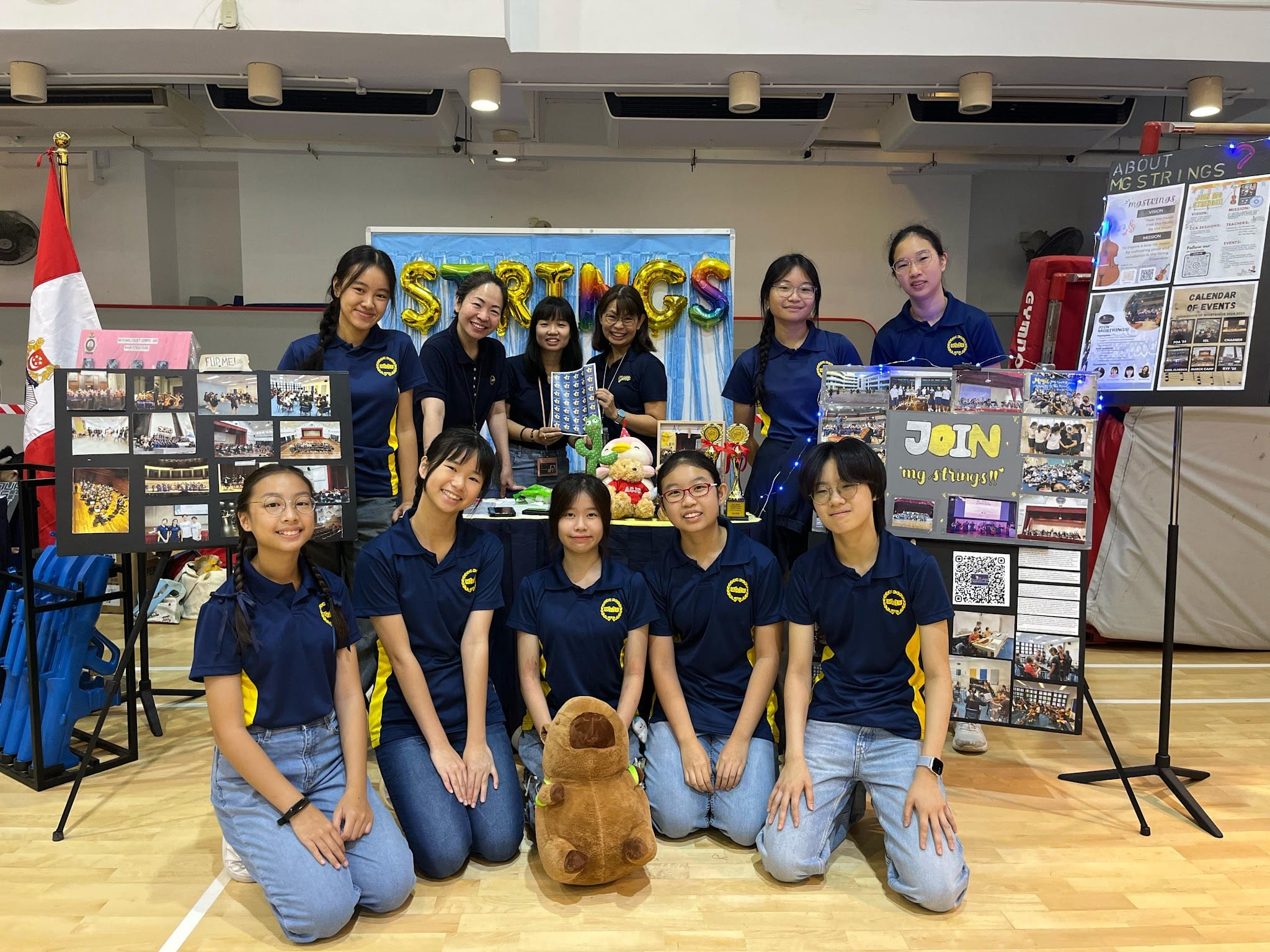 Group of students in navy shirts pose at a school strings club recruitment booth with balloon letters and display boards.