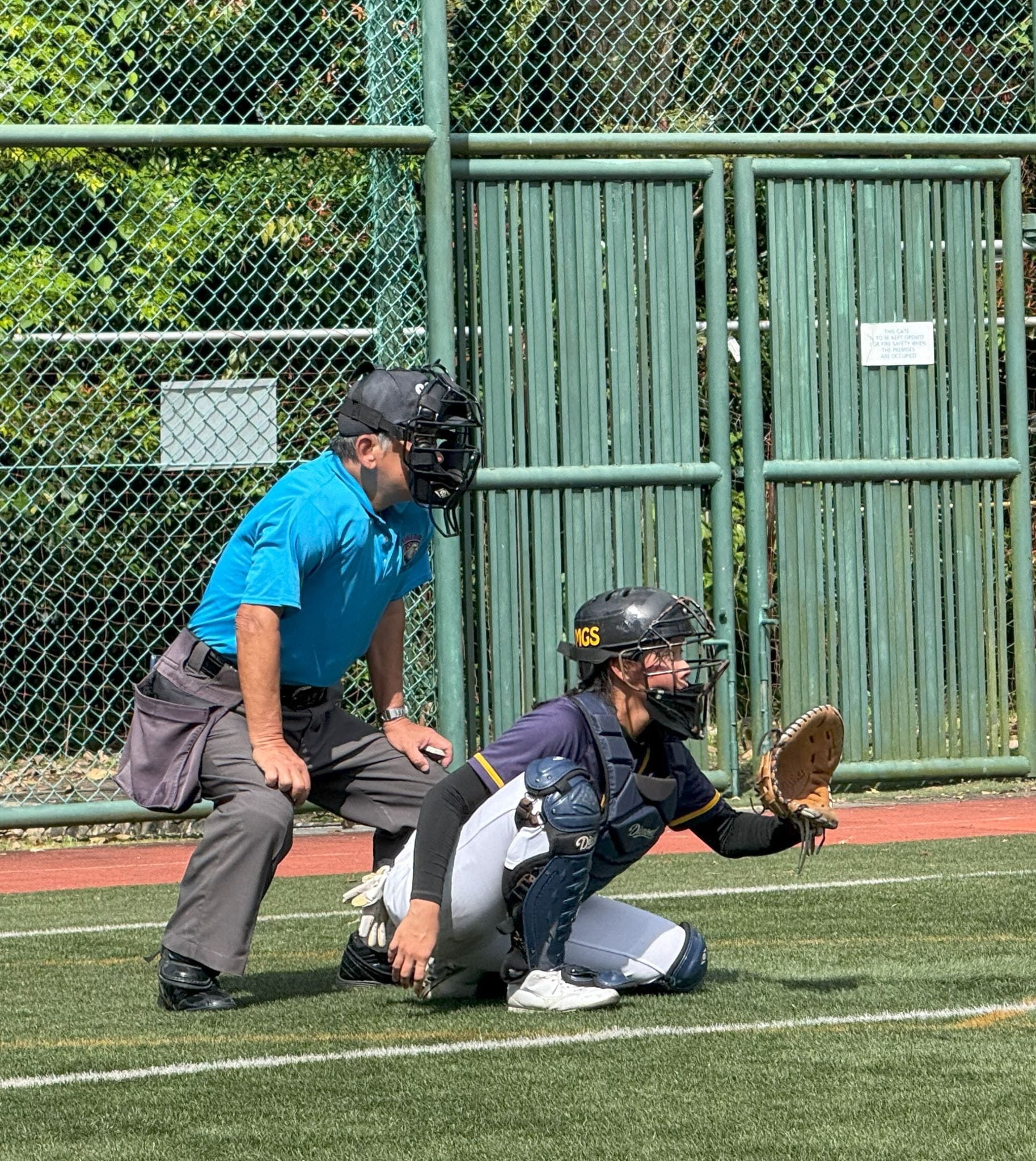A baseball catcher in navy uniform crouches with mitt raised while an umpire in blue shirt stands behind.