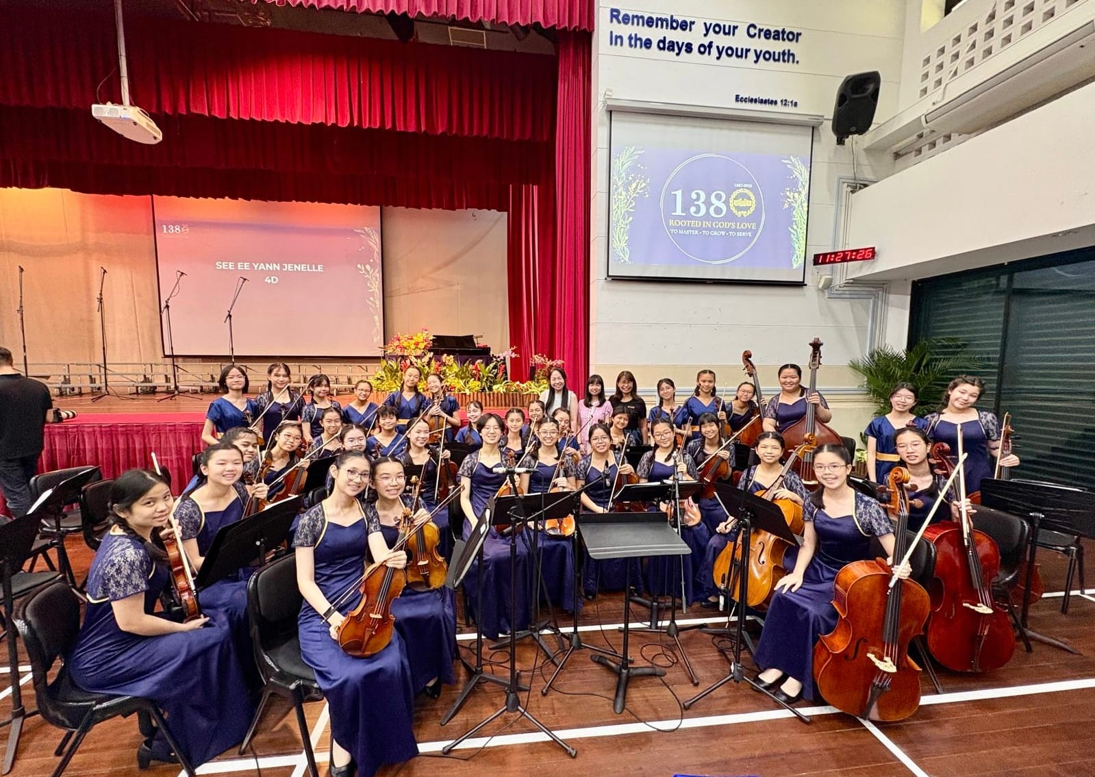 A large youth string orchestra in navy blue dresses posing with violins and cellos in a school hall.