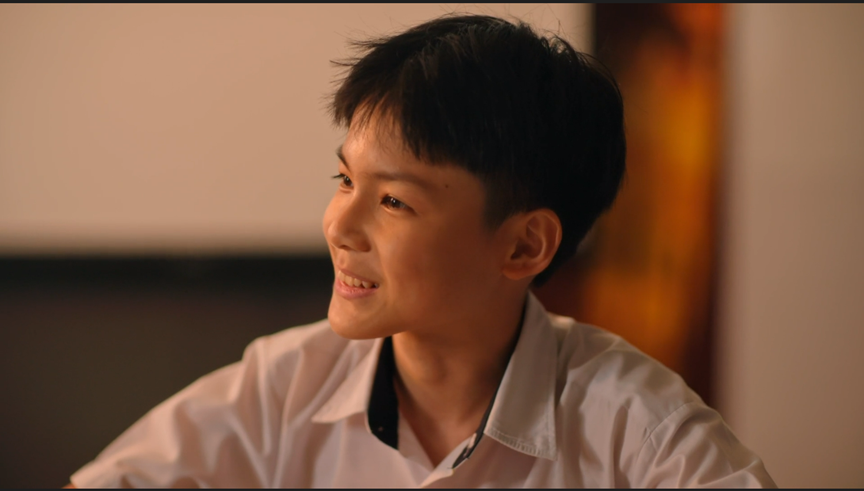 A smiling young boy with short black hair wearing a white collared shirt, looking to the side.