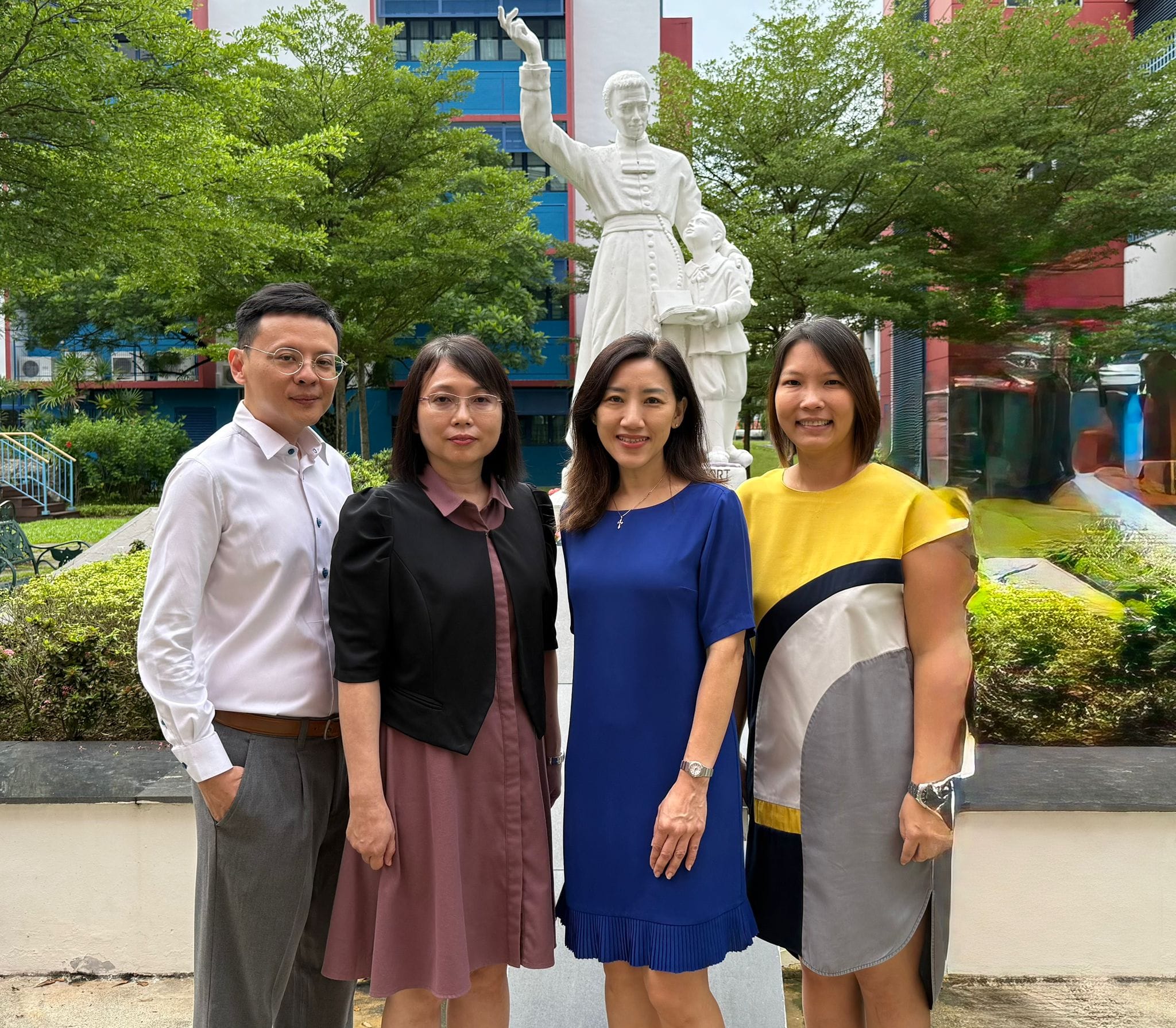 Four people pose outdoors in front of a white statue of a robed figure with raised arm, surrounded by trees and buildings.
