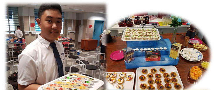 A student in school uniform holds a tray of decorated snacks beside a table displaying various pastries and foods.