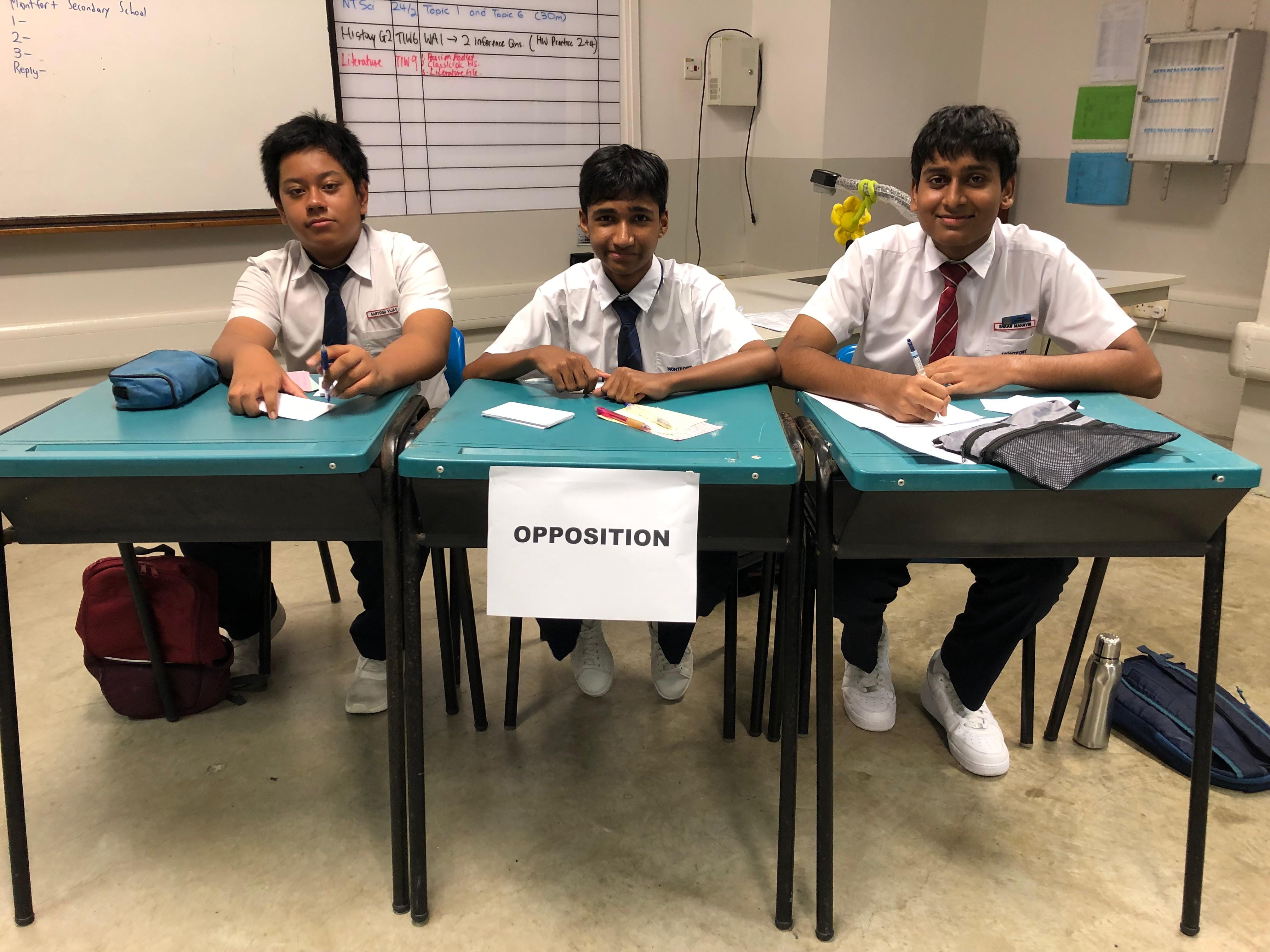 Three male students in school uniforms sit at desks labeled Opposition during a classroom debate.