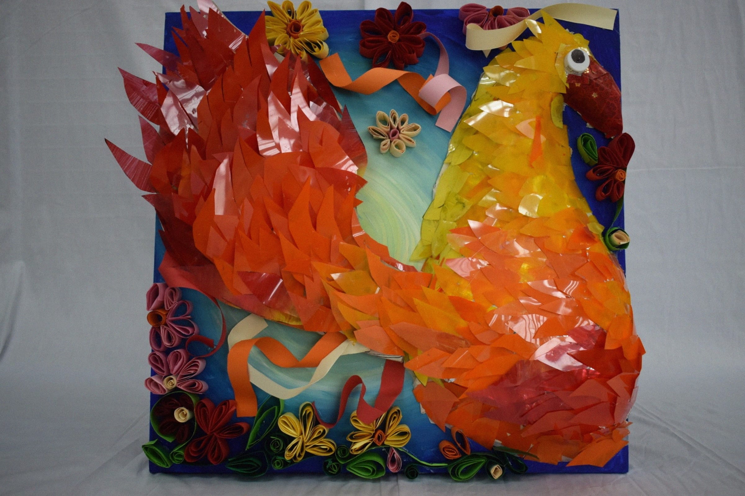A colorful paper collage of a phoenix bird in red, orange, and yellow, surrounded by quilled paper flowers on a blue board.