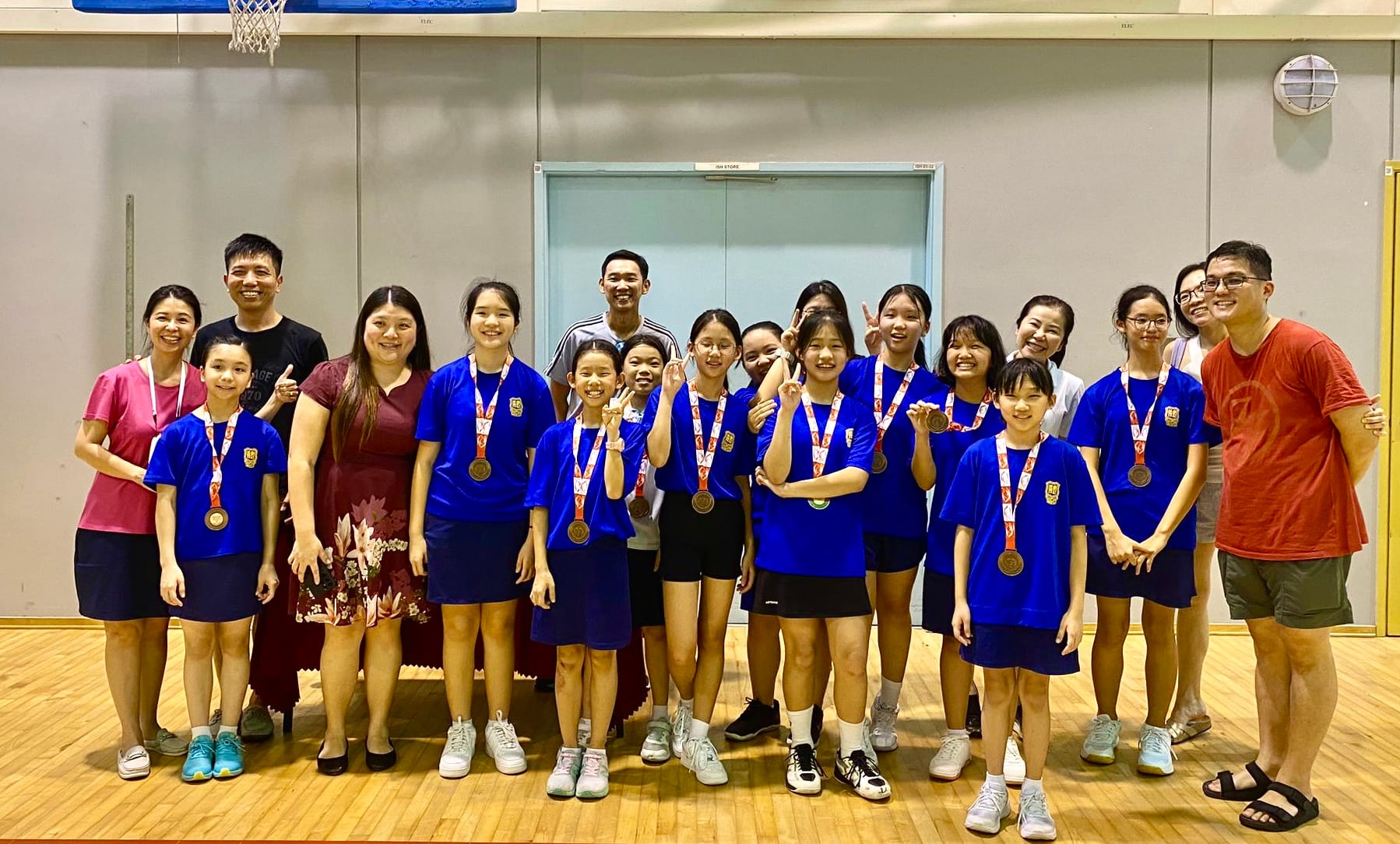 A group of youth in blue shirts wearing bronze medals pose with adults in a gymnasium.