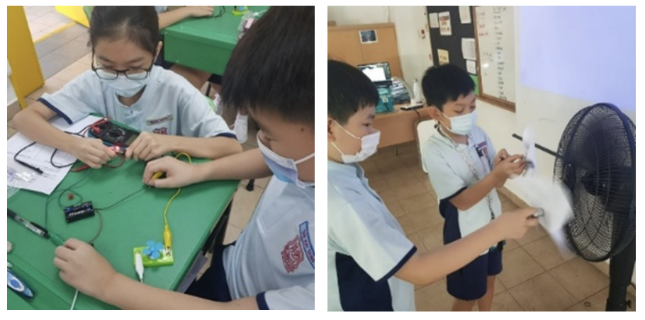 Two side-by-side photos of primary school students wearing face masks and school uniforms participating in hands-on science experiments. In the image on the left, a boy and a girl sit at a green desk working with an electrical circuit kit that includes wires, a battery pack, a multimeter, and a small fan with blue blades. In the image on the right, two boys stand in a classroom testing an object in front of a running black electric floor fan.