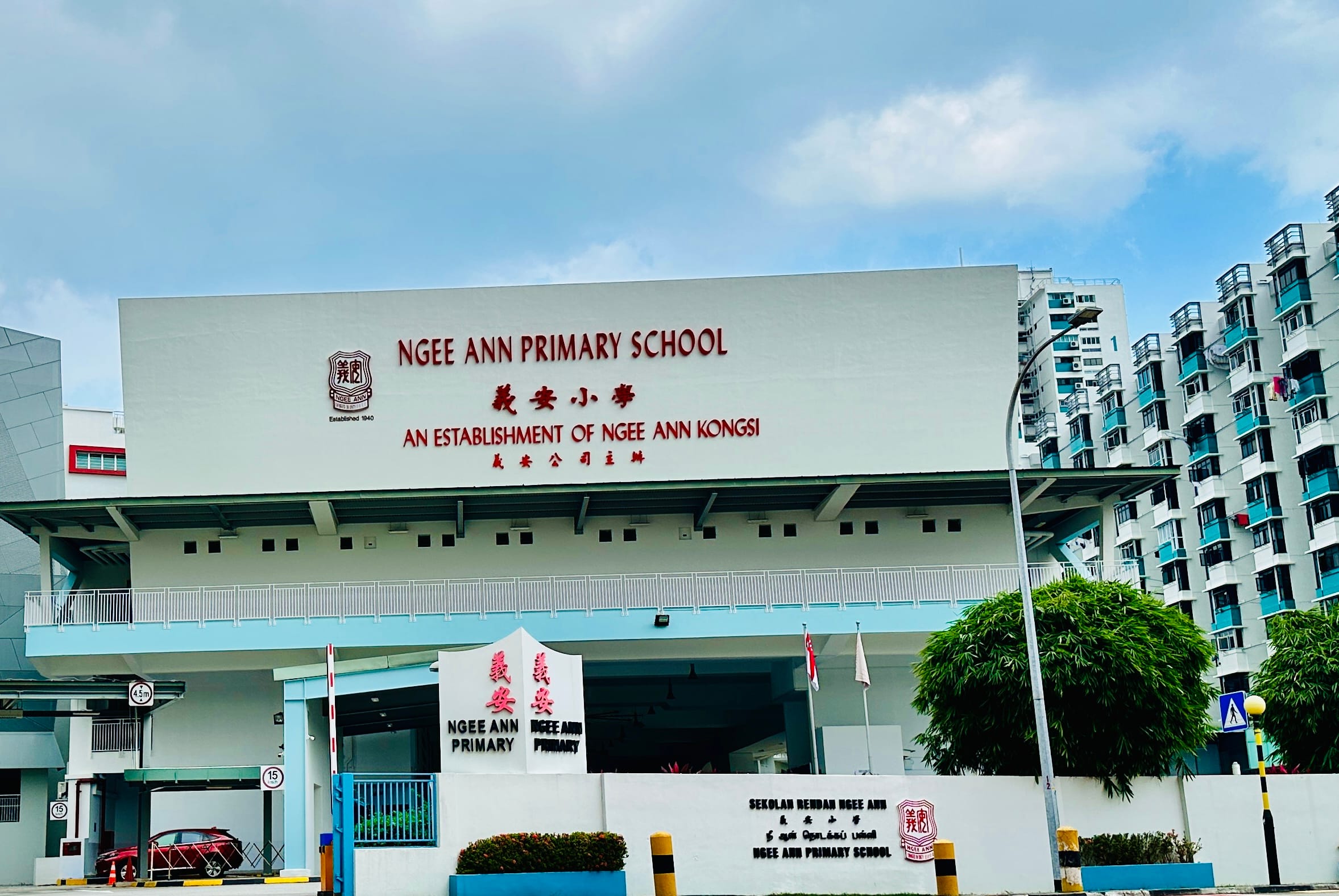 Exterior of Ngee Ann Primary School building with red signage in English and Chinese, flanked by residential buildings.
