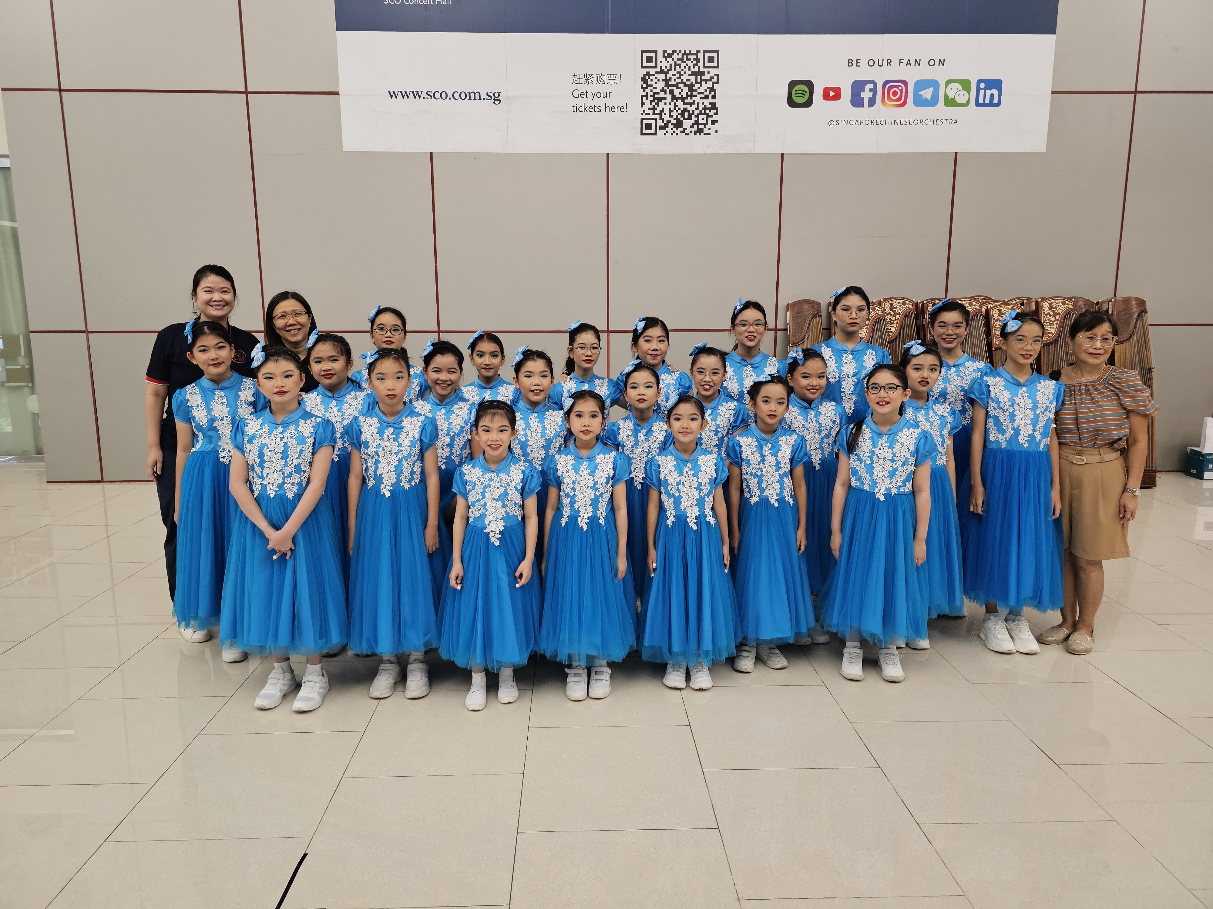 A group of young girls in matching blue dresses with white lace pose with adults in front of an SCO banner.