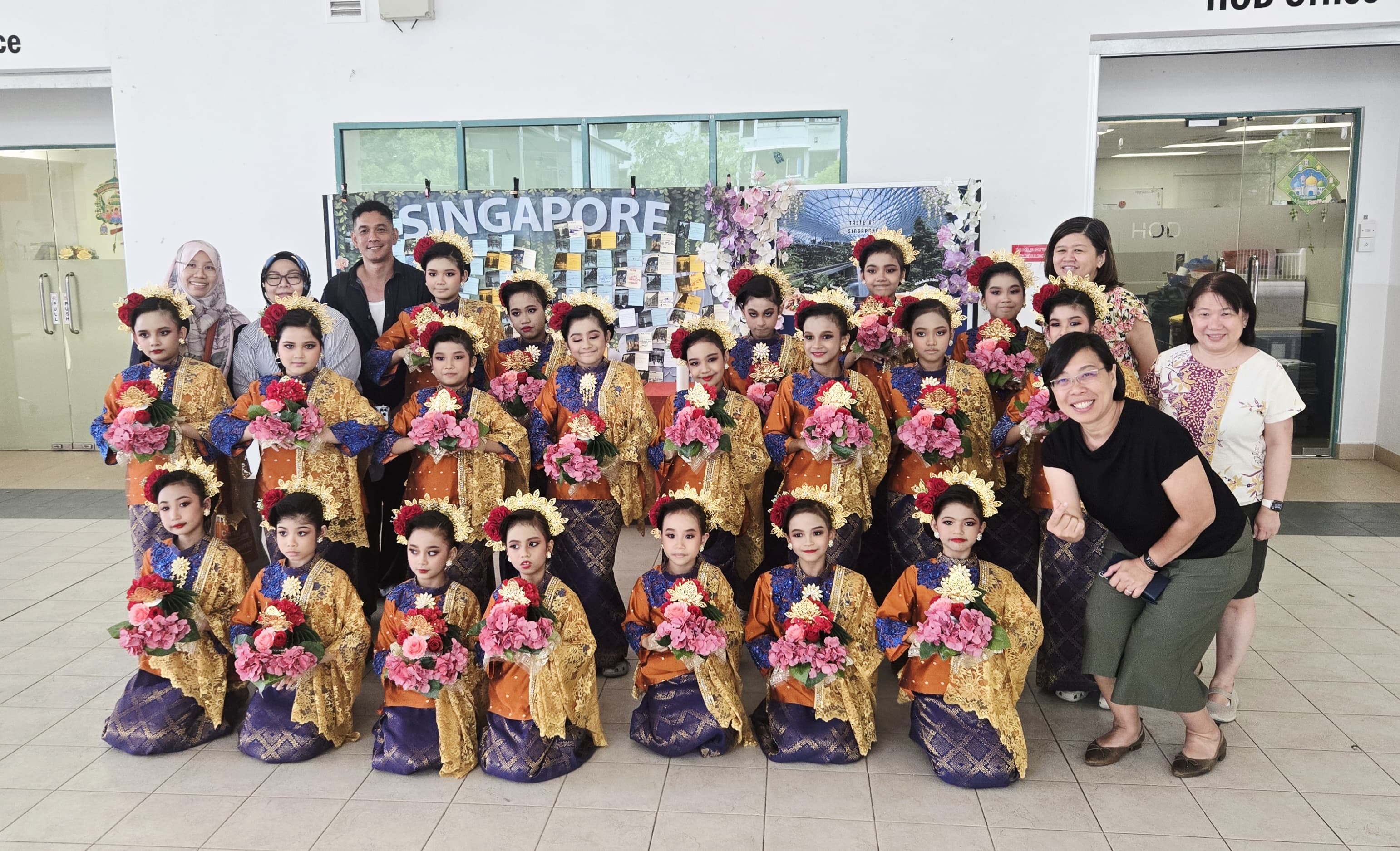 Group of children in traditional Malay gold and orange costumes holding flowers pose with adults before a Singapore display board.