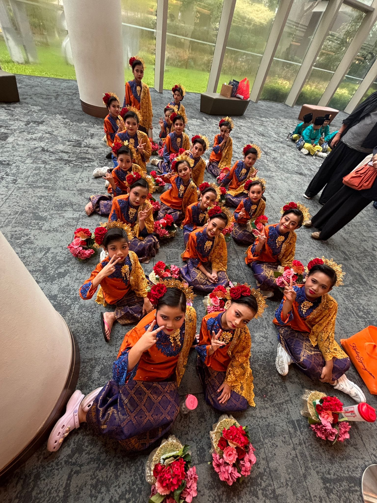 A group of children in orange and purple traditional costumes with floral headpieces sit on a floor posing with peace signs.