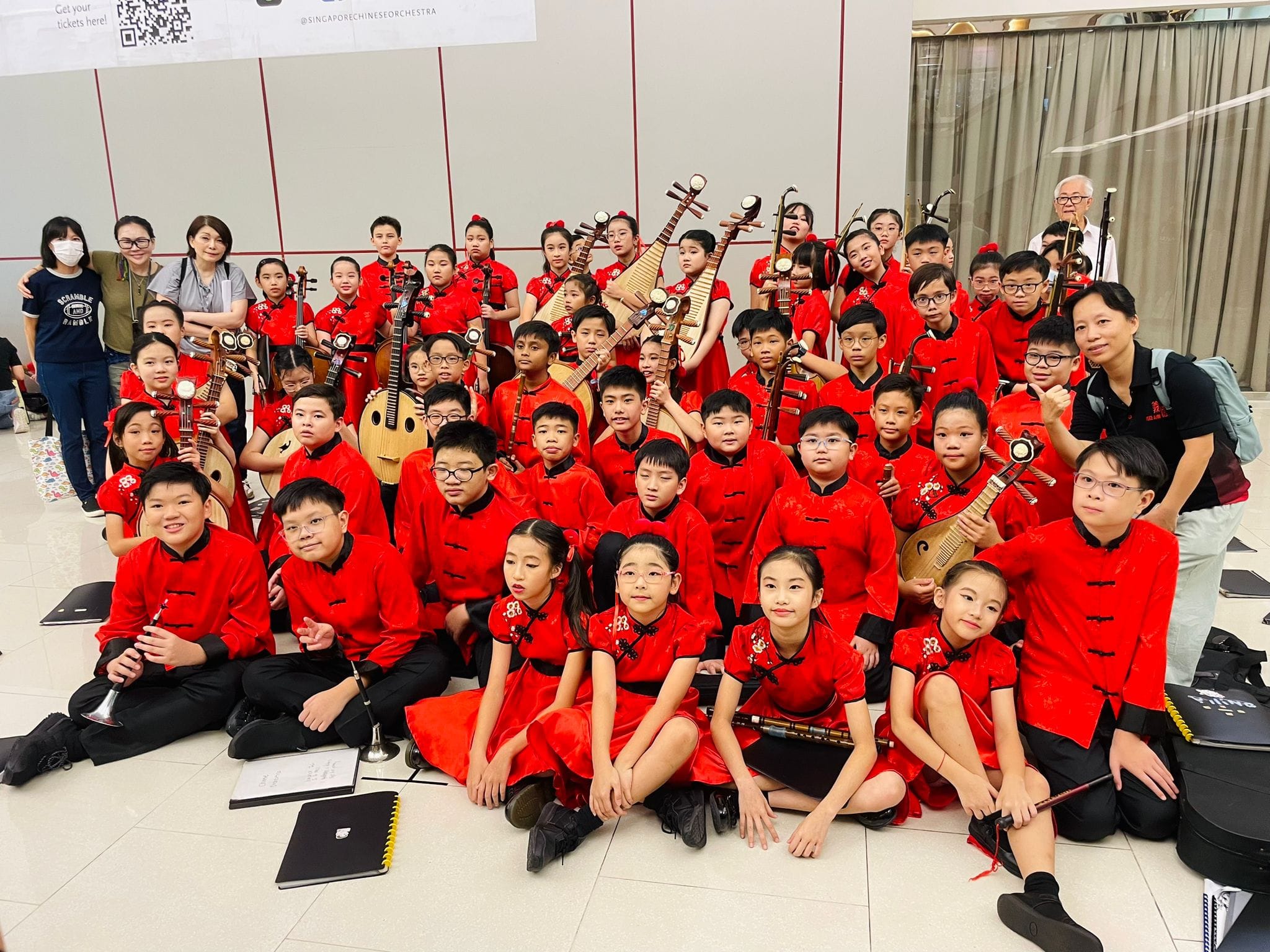 A large group of children in red traditional Chinese outfits pose together holding Chinese instruments like erhu and pipa.