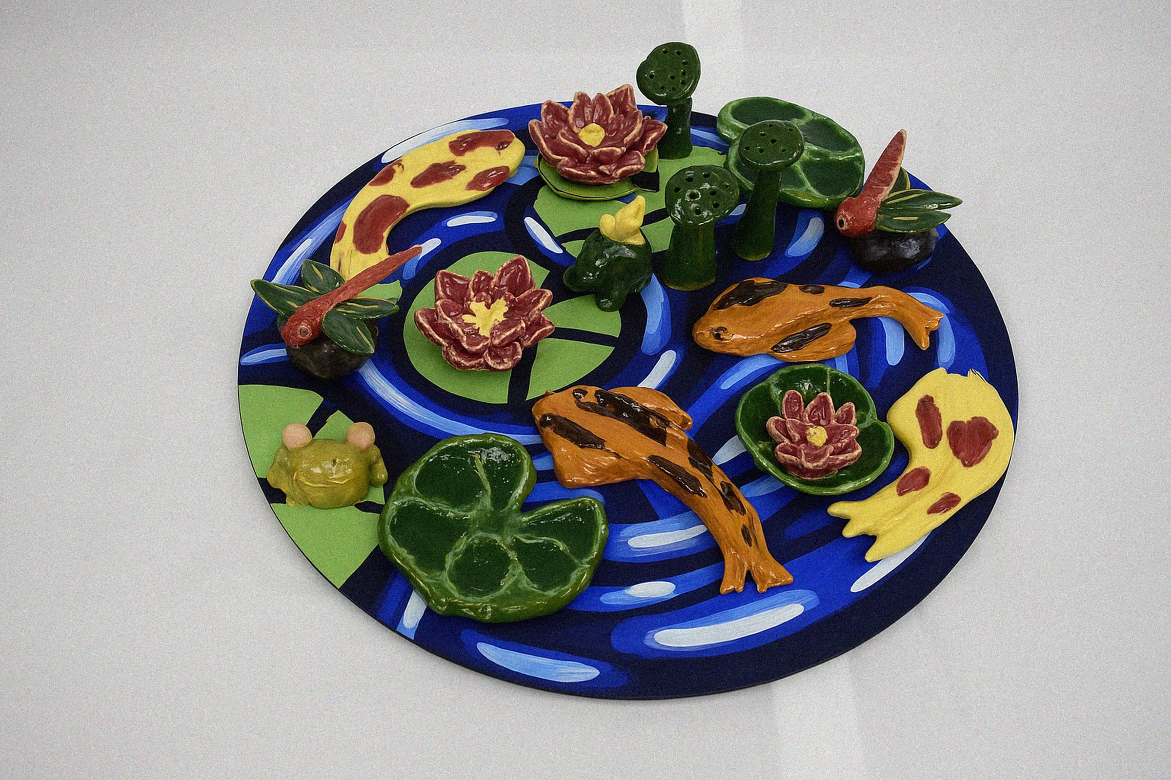 Round painted pond sculpture with lily pads, koi fish, a frog, flowers, and small green trees.