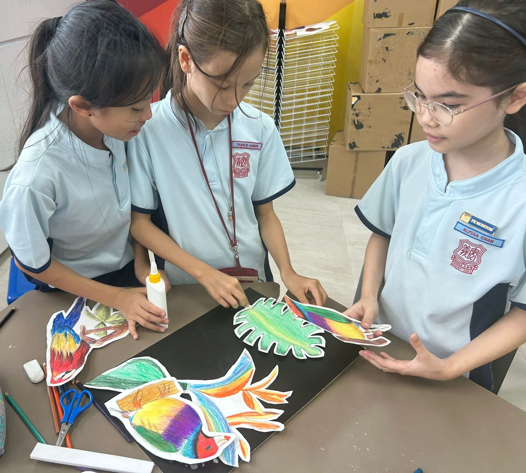 Three schoolgirls in uniforms arrange colorful cut-out bird drawings on black paper using glue.