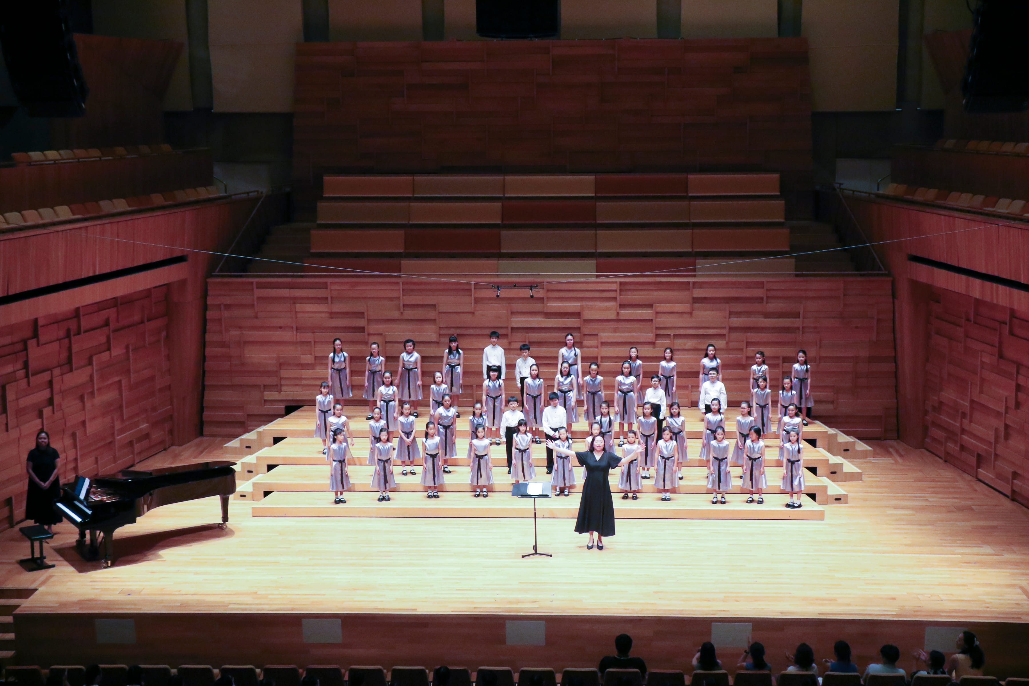 A conductor in black leads a large children's choir on a wooden concert hall stage, with a pianist at a grand piano nearby.