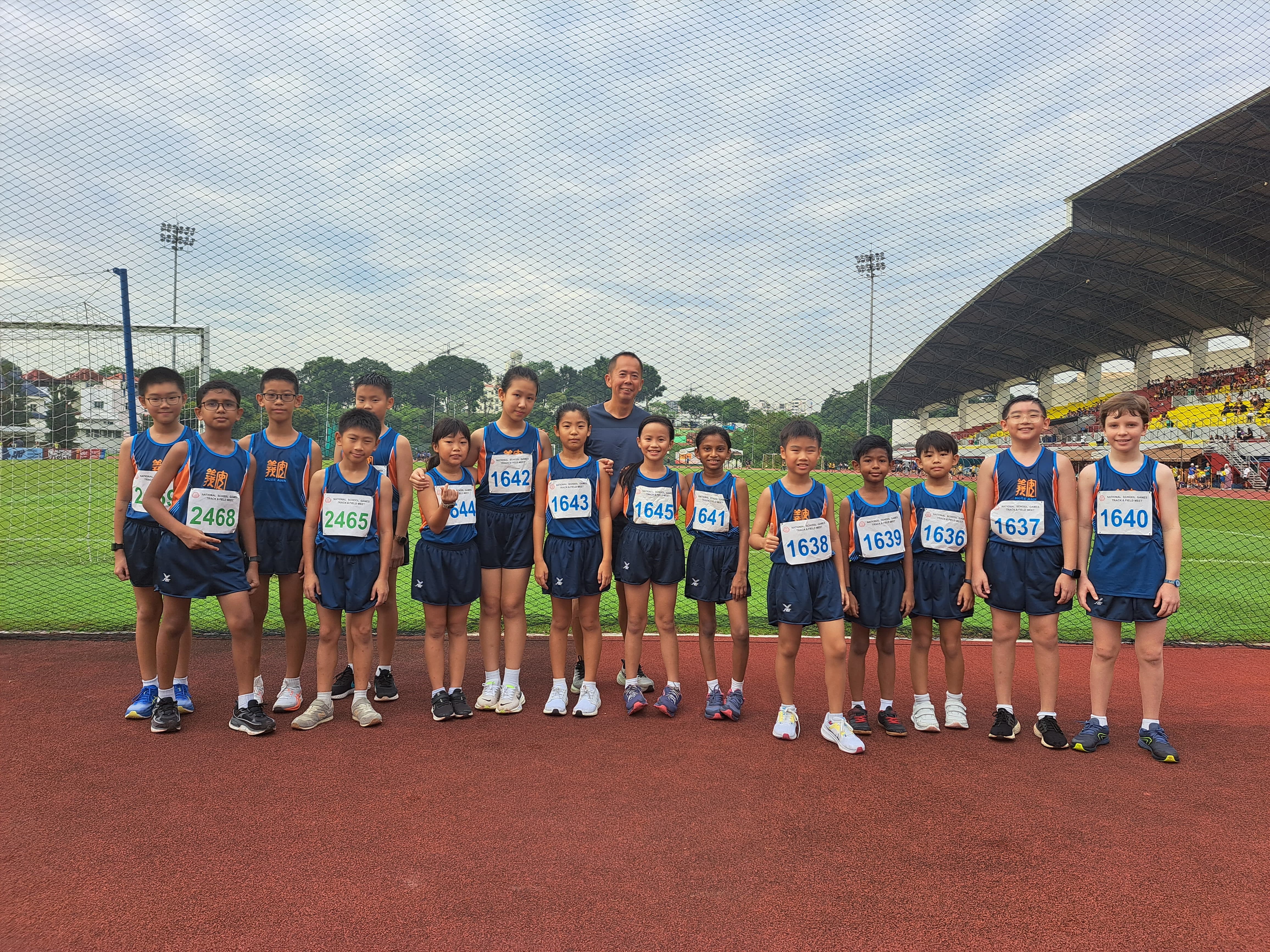 Group of young athletes in blue uniforms with race bibs posing with a coach on a red running track at a stadium.
