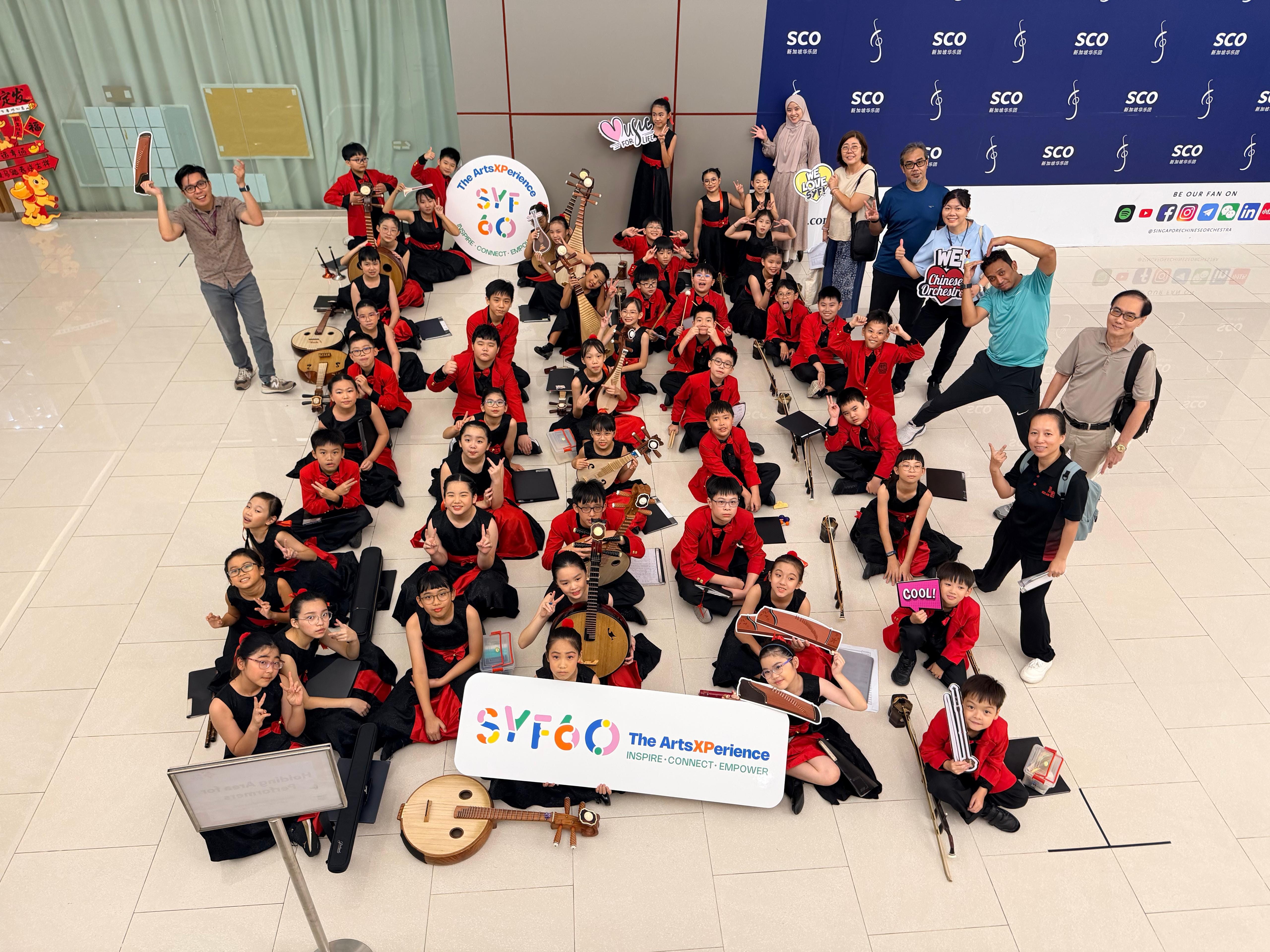 Large group of children in red and black uniforms holding Chinese instruments, posing with adults at an SCO arts event.