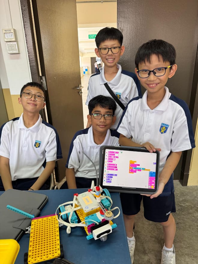 Five Ngee Ann Secondary students posing with their LEGO robot and iPad displaying block-coding software.