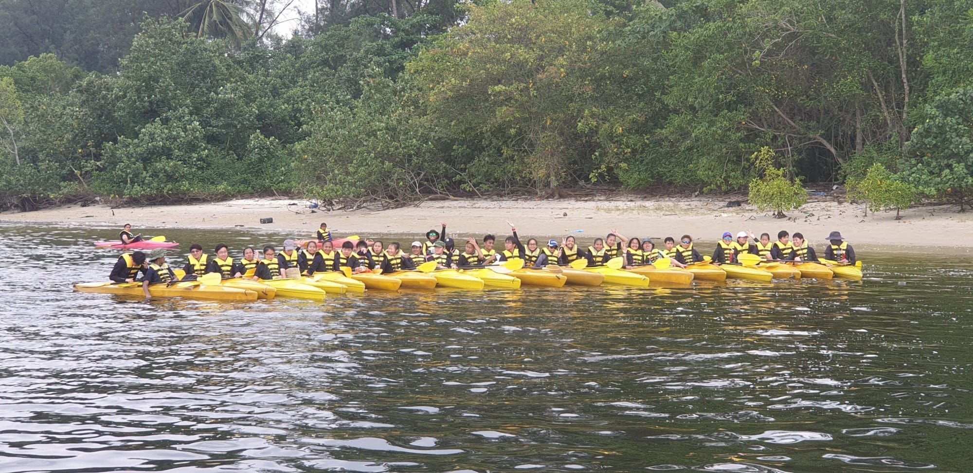 Small groups of Ngee Ann Secondary students wearing yellow life vests sitting in yellow kayaks which are lined up near the shore.