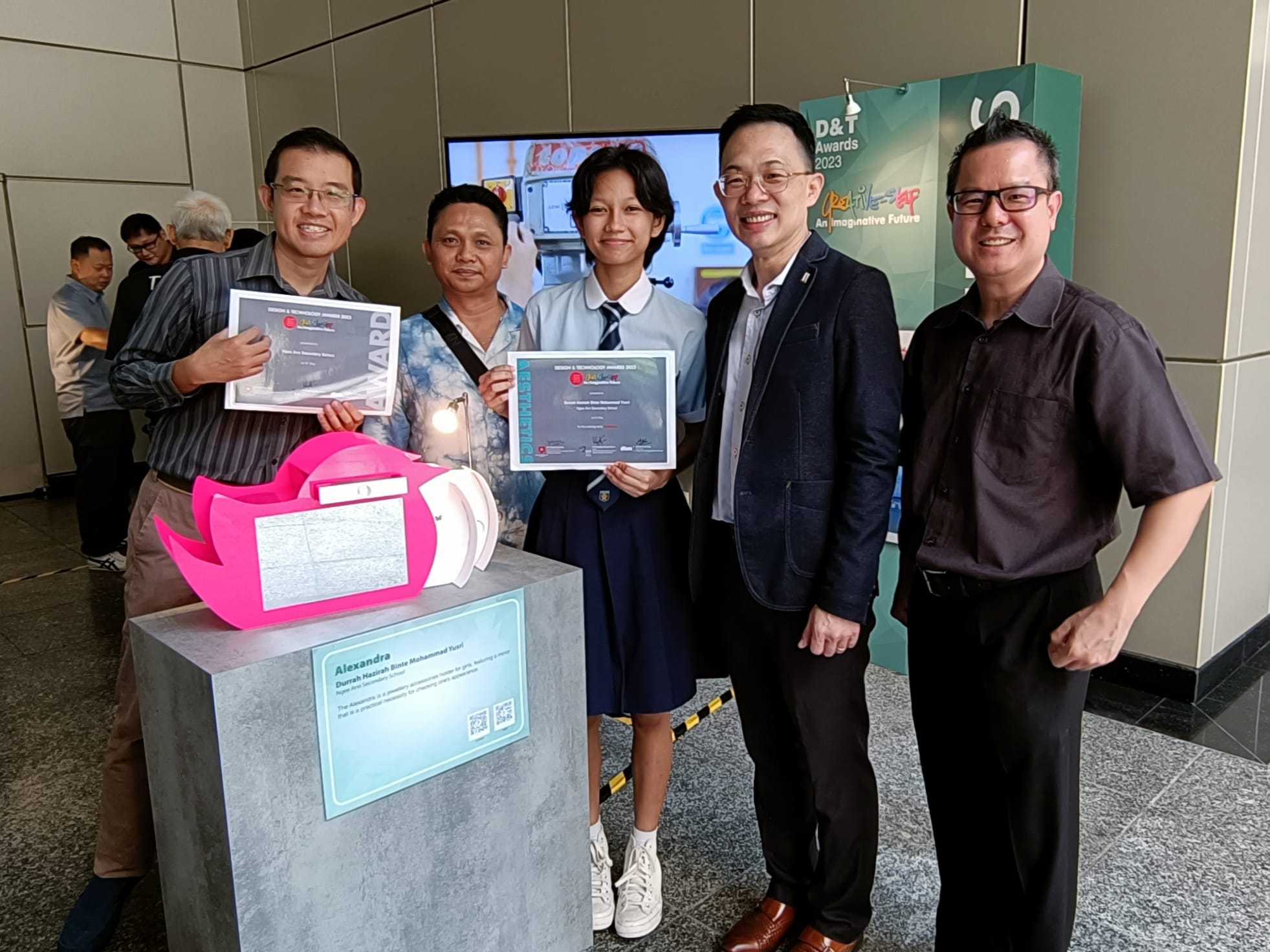 Five people posing at a D&T Awards 2023 event, holding certificates while standing next to a student's project.