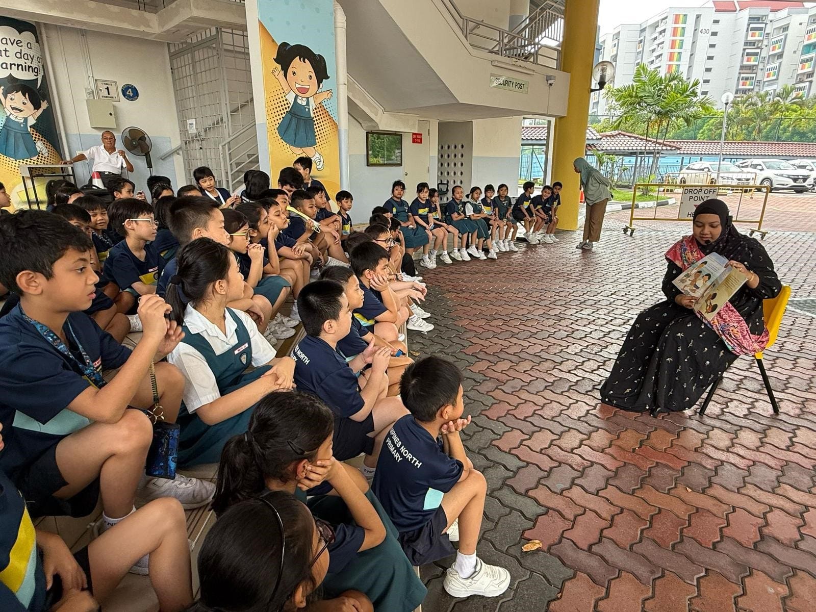A woman in black reads a book to a large group of uniformed primary school children seated outdoors.