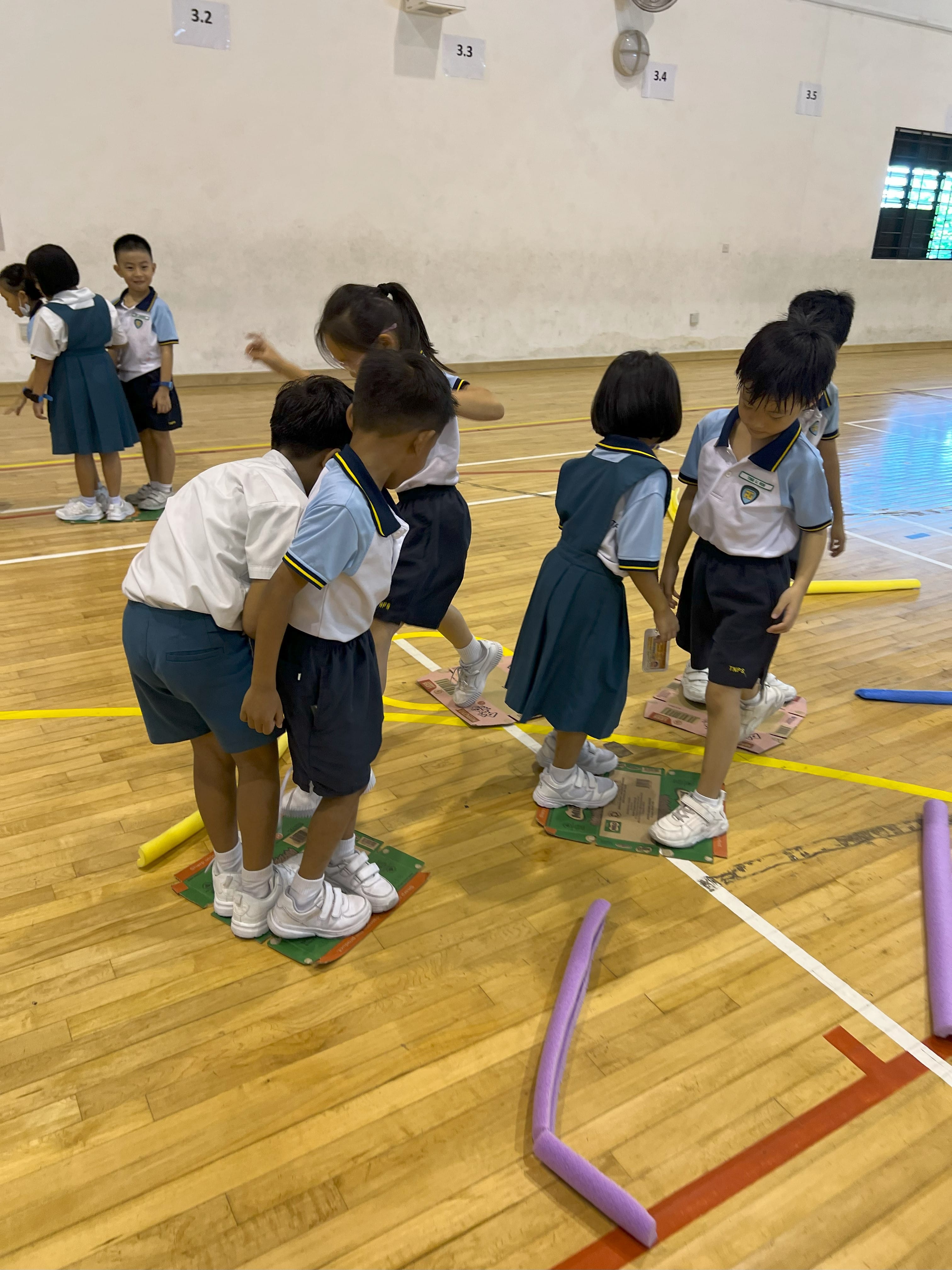 Young students in school uniforms stand on cardboard pieces during a gym activity on a wooden floor.