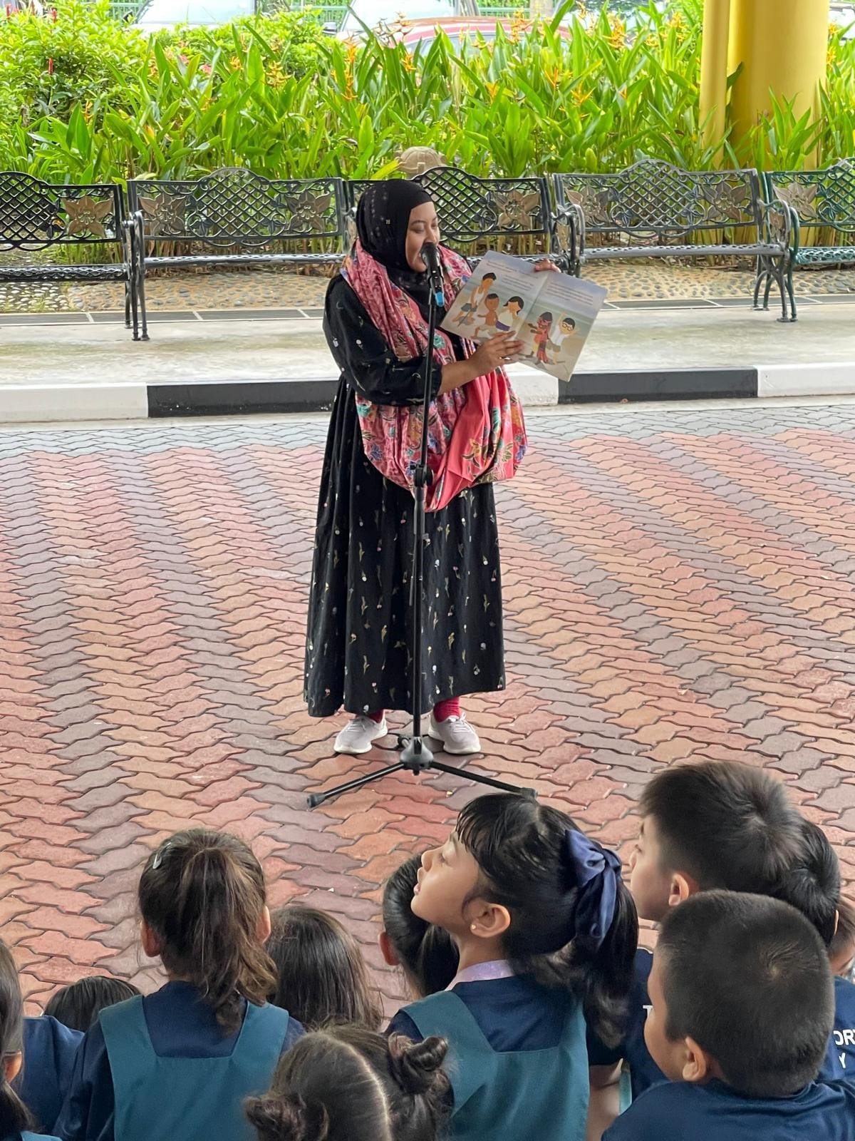 A woman in a black hijab reads a picture book aloud at a microphone to a group of young children outdoors.