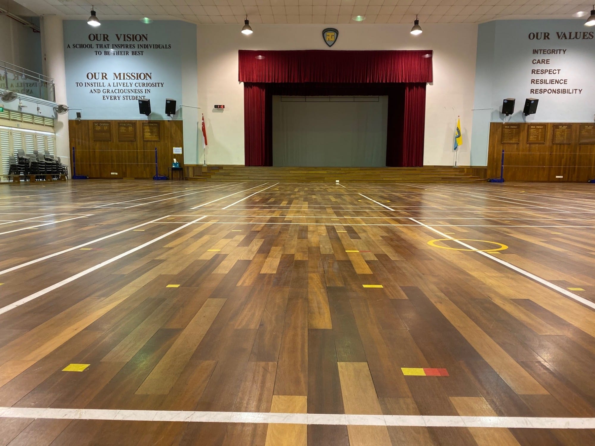 Empty school hall with wooden floor, a red-curtained stage, and walls displaying the school's vision, mission, and values.