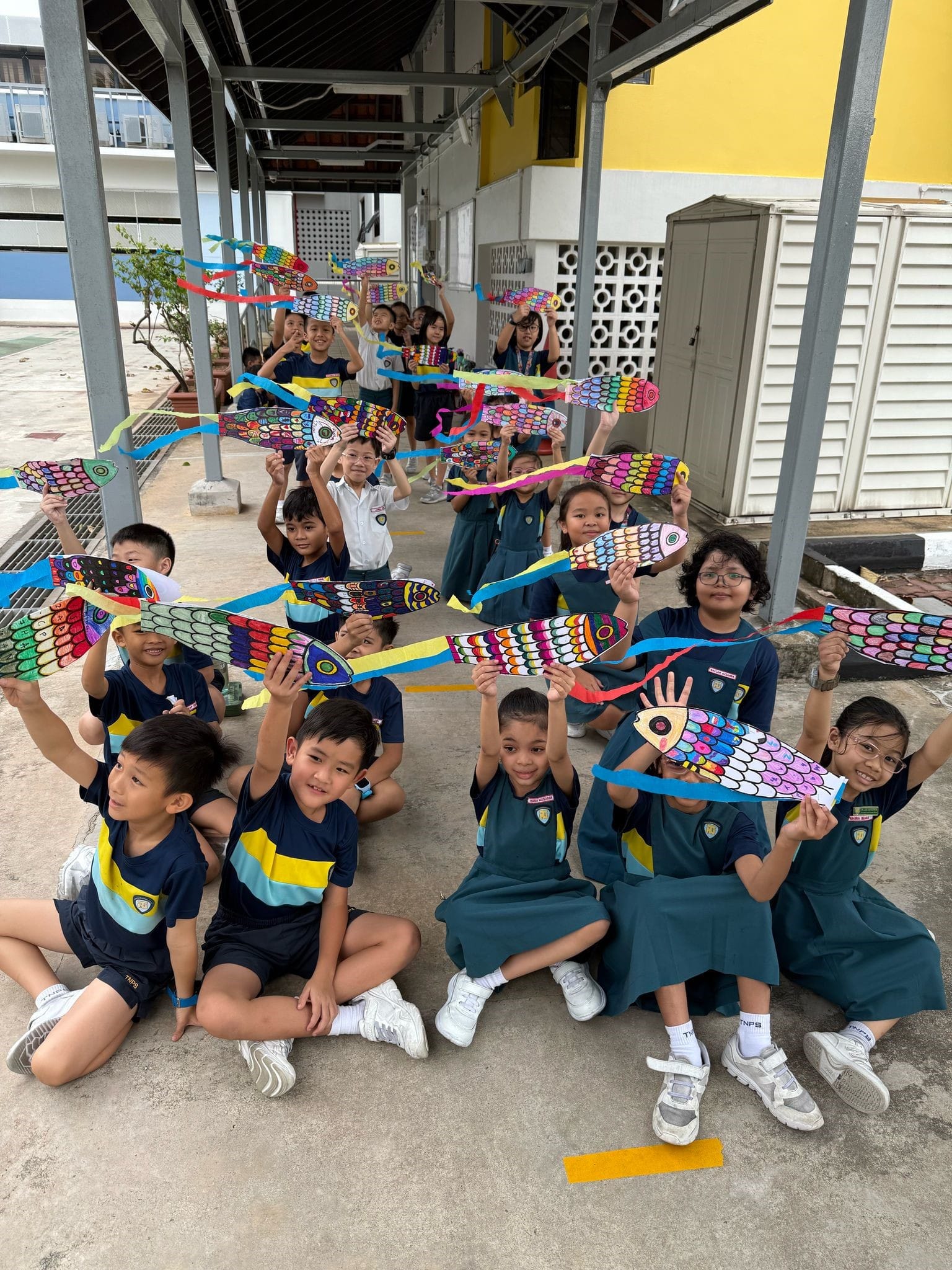 A group of school children in uniforms holding up colorful hand-decorated paper fish kites outdoors.