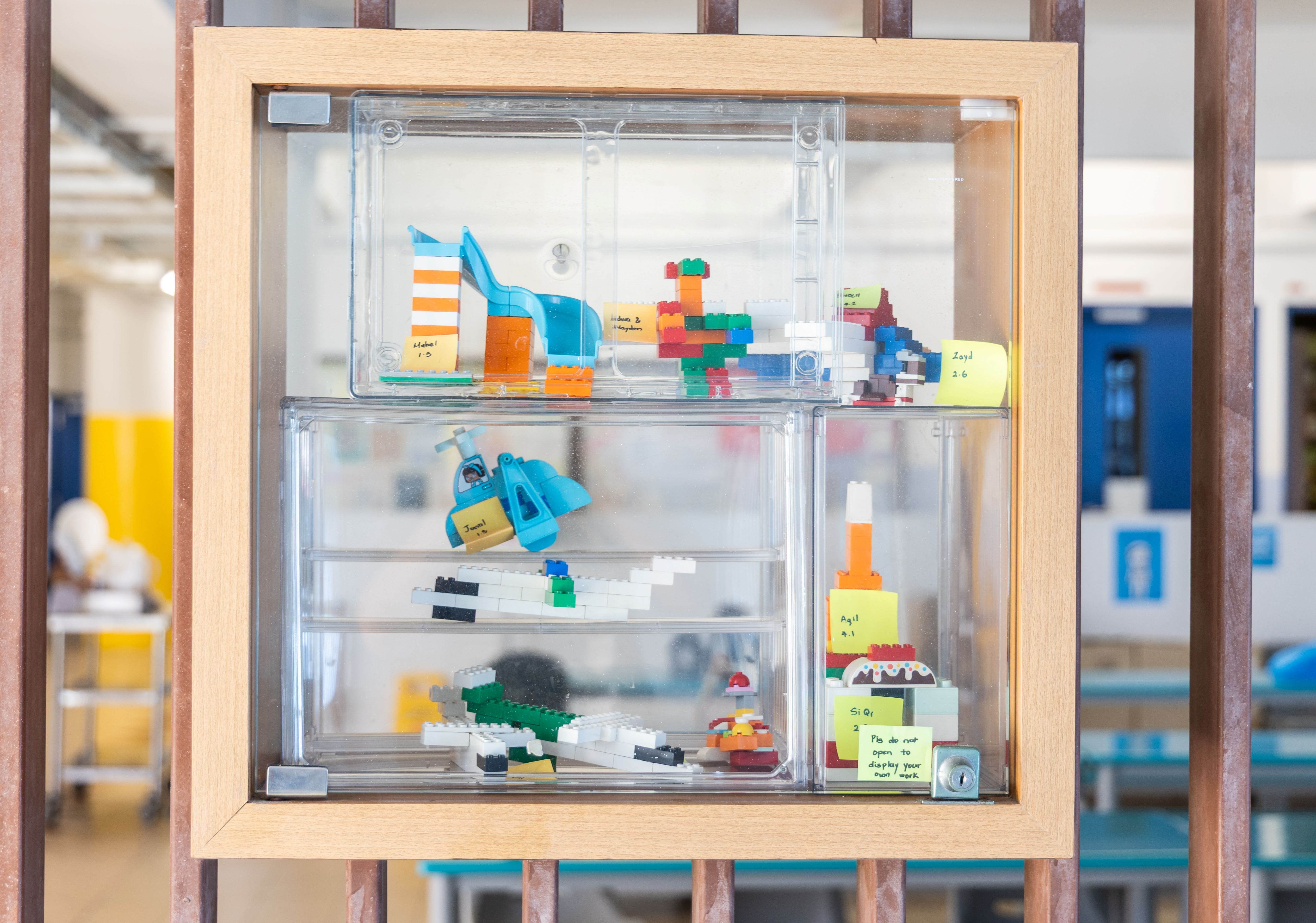 Wooden display case with clear compartments holding colorful LEGO builds and small yellow sticky note labels.