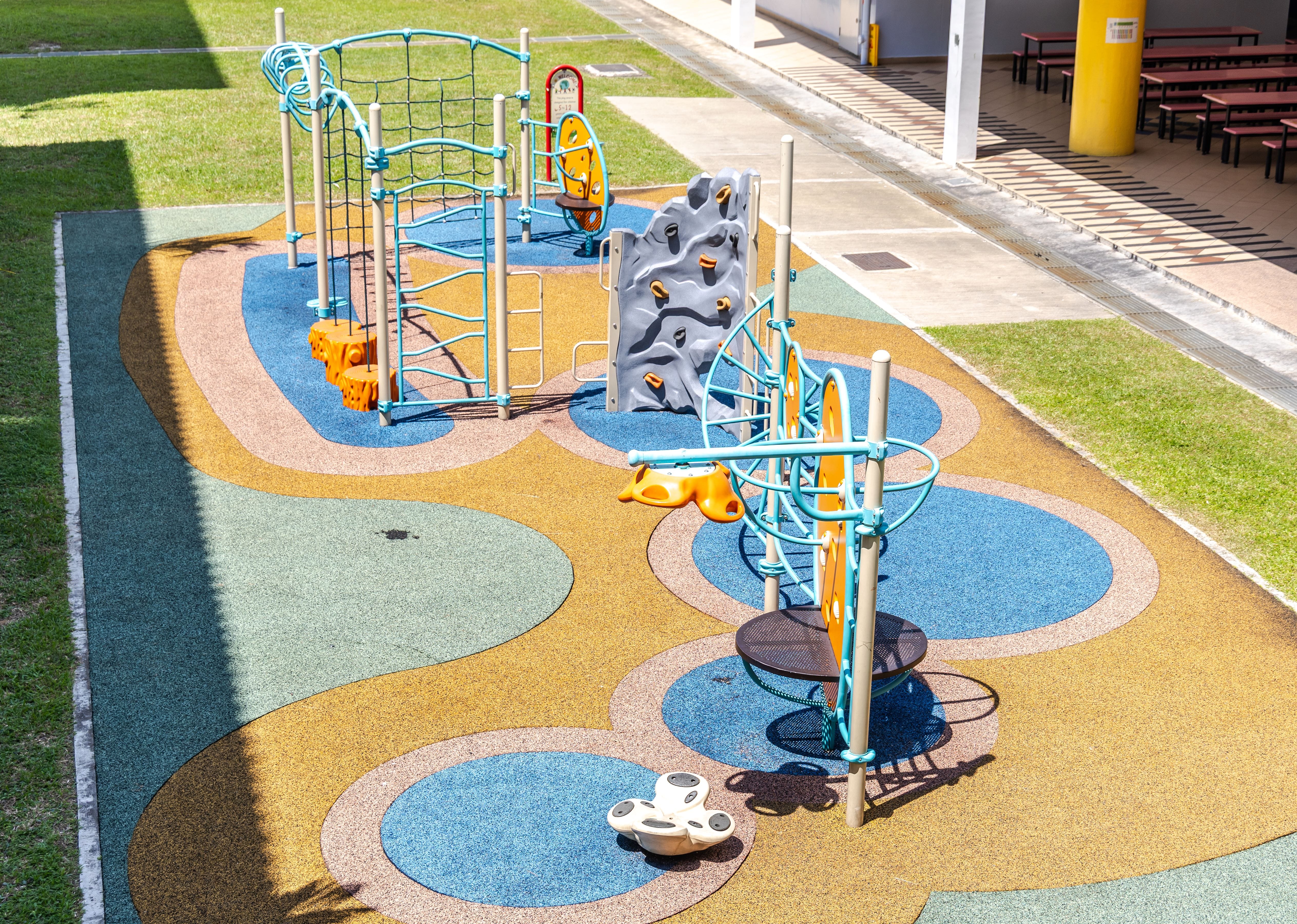 Empty playground with climbing net, rock wall, and spinner on colorful rubber surface beside grass and tables