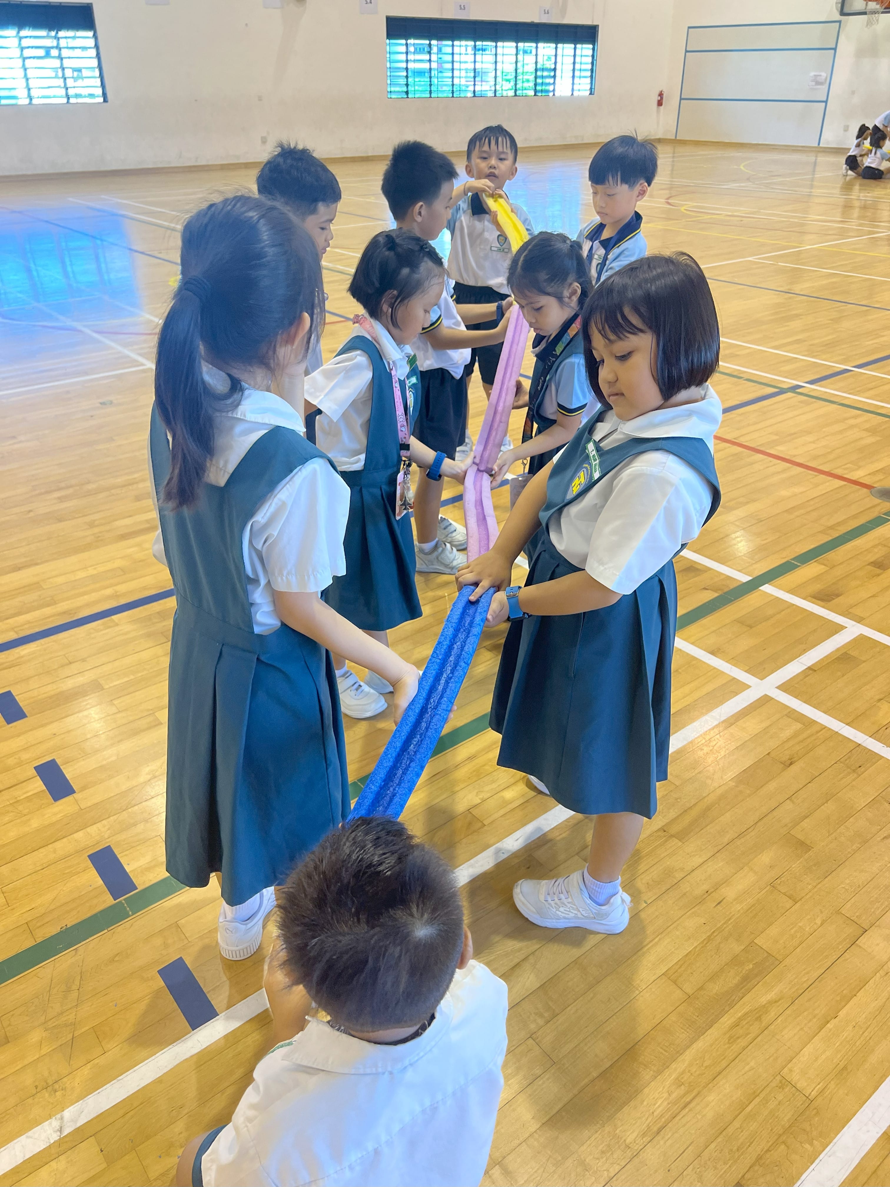 Young schoolchildren in uniforms hold colorful fabric strips together in a group activity on a gymnasium floor.
