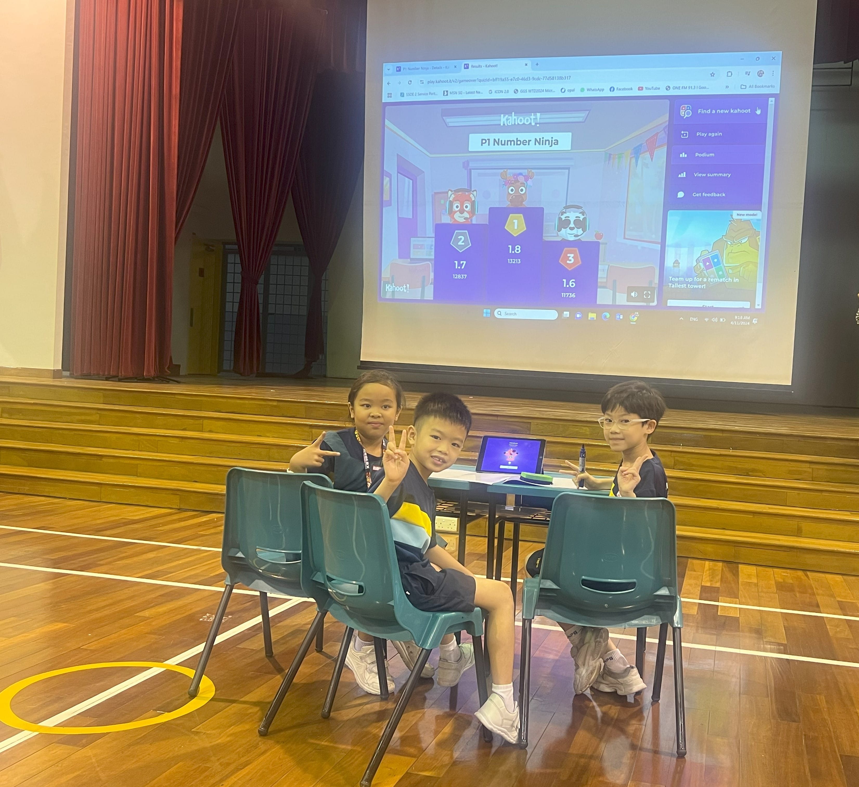 Three young children smile and pose at a table in a school hall with a Kahoot results screen projected behind them.